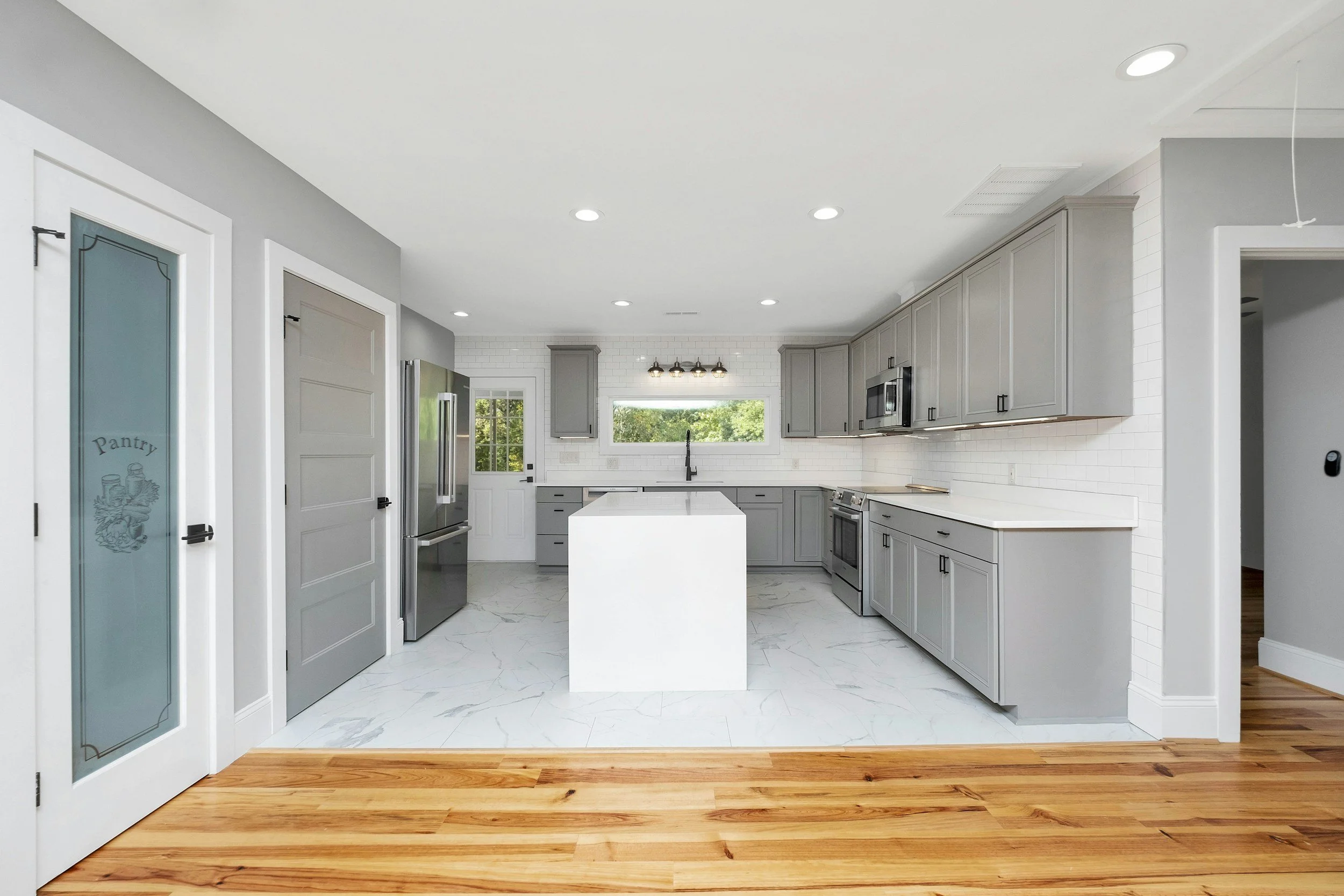 Modern kitchen with white countertops, gray cabinets, stainless steel appliances, and a centrally placed white island. There is a window above the sink and a small pantry door on the left with a frosted glass panel.