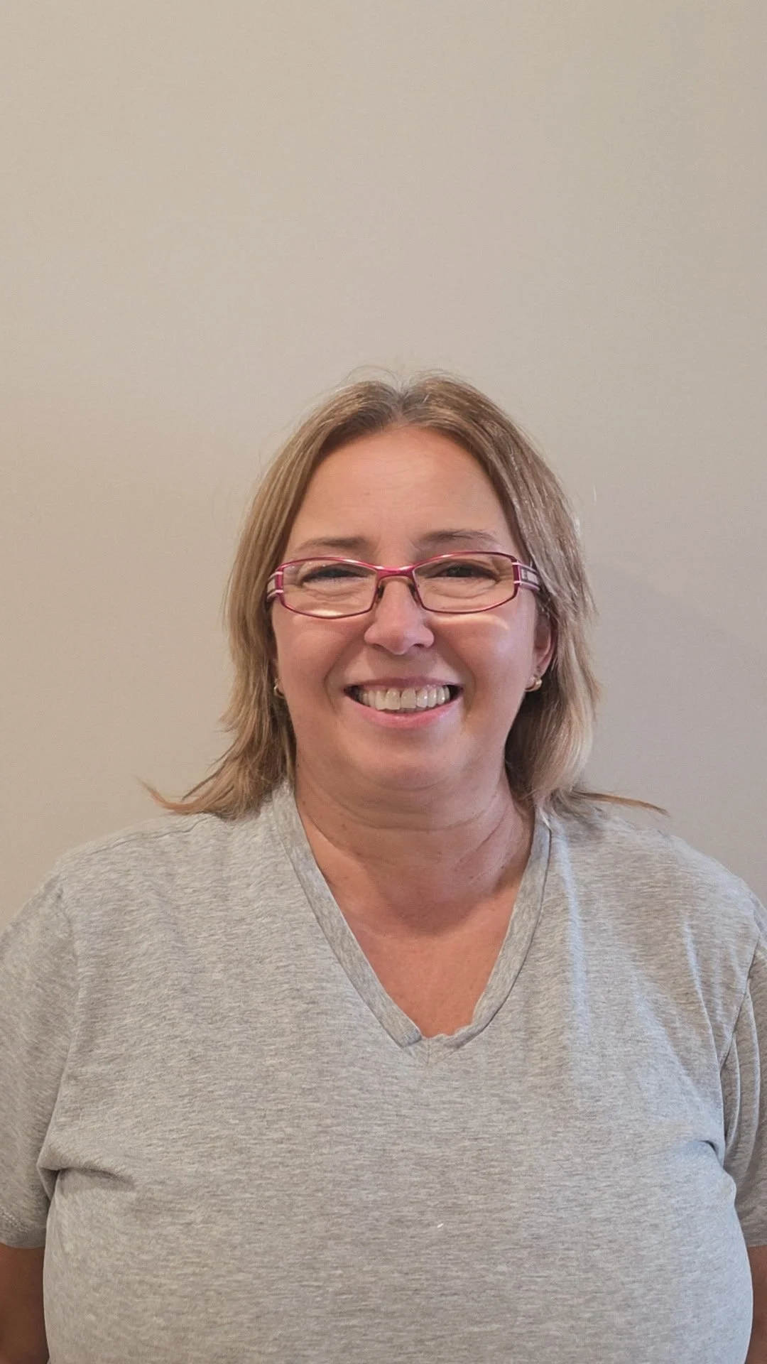 A smiling woman with shoulder-length blonde hair, wearing glasses and a light gray t-shirt, standing against a plain beige background.