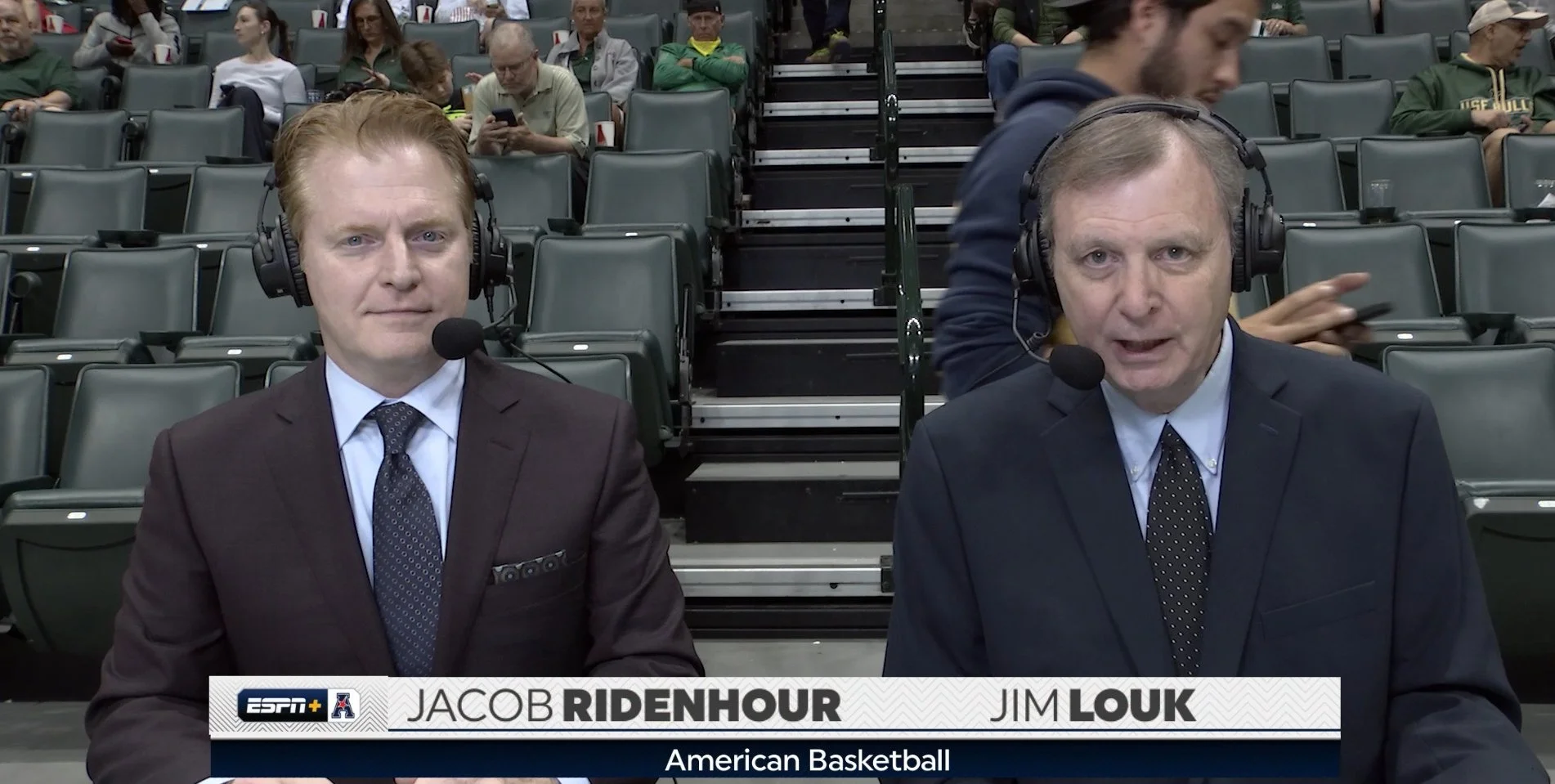 Two men in suits with headsets and microphones sitting at a sports broadcast table, with empty stadium seats and some audience members in the background, reporting on American basketball.
