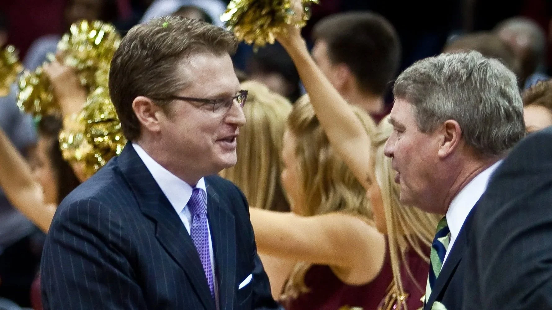 Two men in suits engaging in conversation at a gathering, with cheerleaders holding gold pom-poms in the background.
