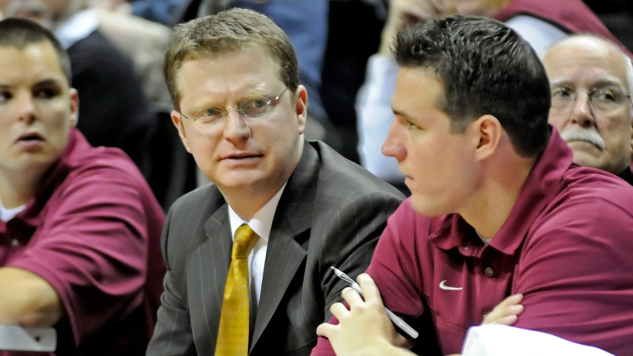Three men sitting at a table in a serious discussion, with the man on the right taking notes, in a formal setting.