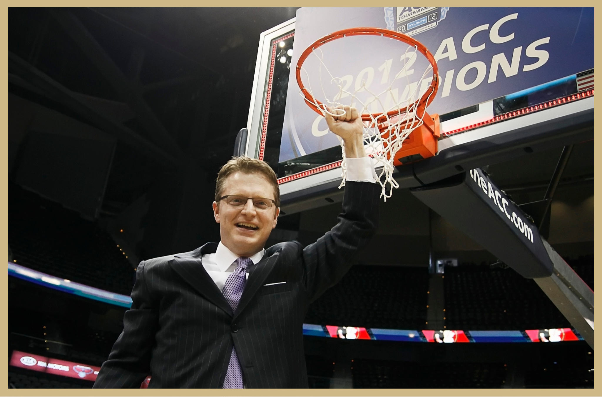 A man in a suit with glasses and a purple tie is smiling and holding up a basketball net after a game, in an indoor arena with a basketball hoop and scoreboard.