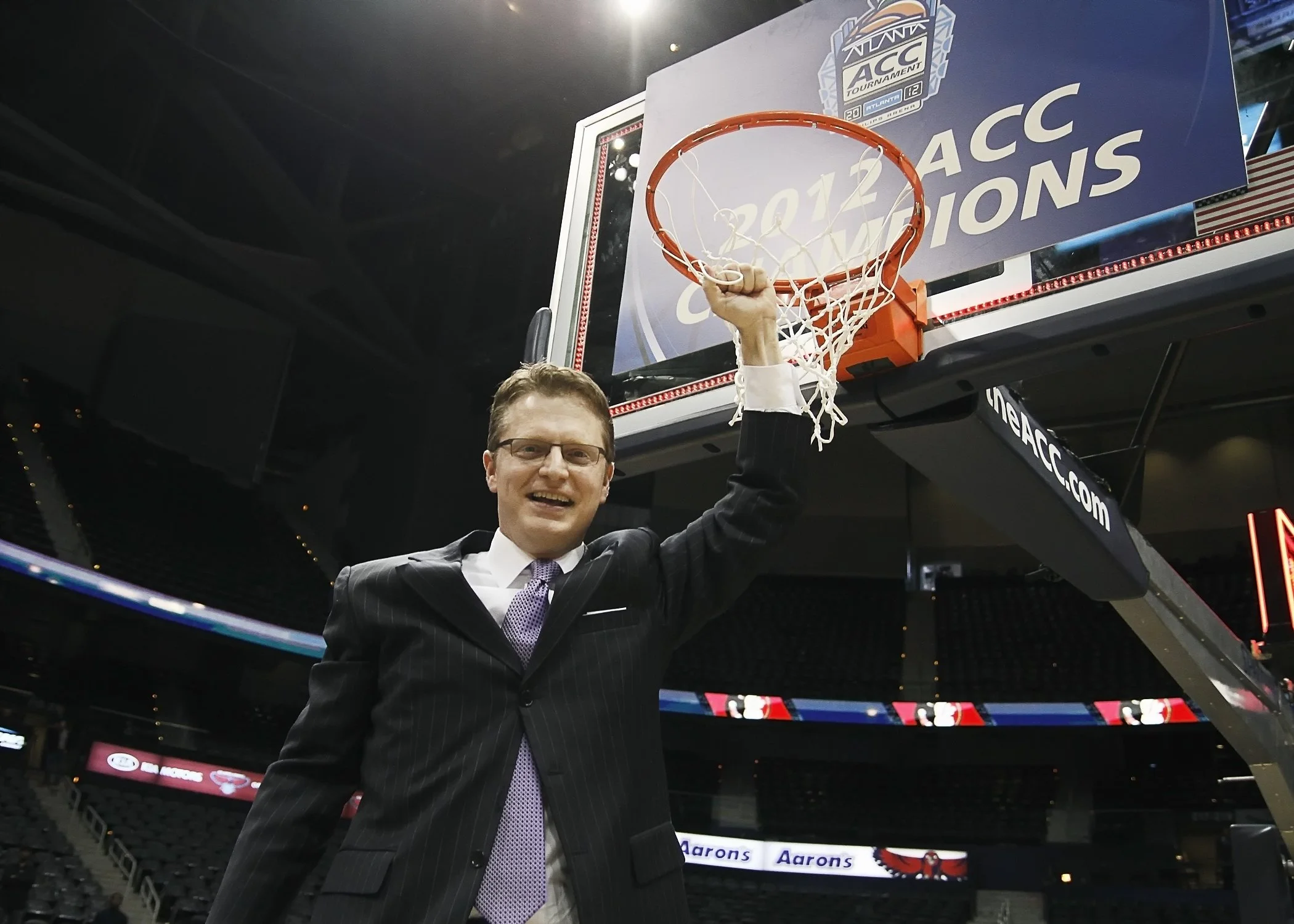 A man in a suit and tie is smiling and raising a basketball hoop with his hand inside a sports arena, with a large banner that reads '2012 ACC CHAMPIONS' in the background.