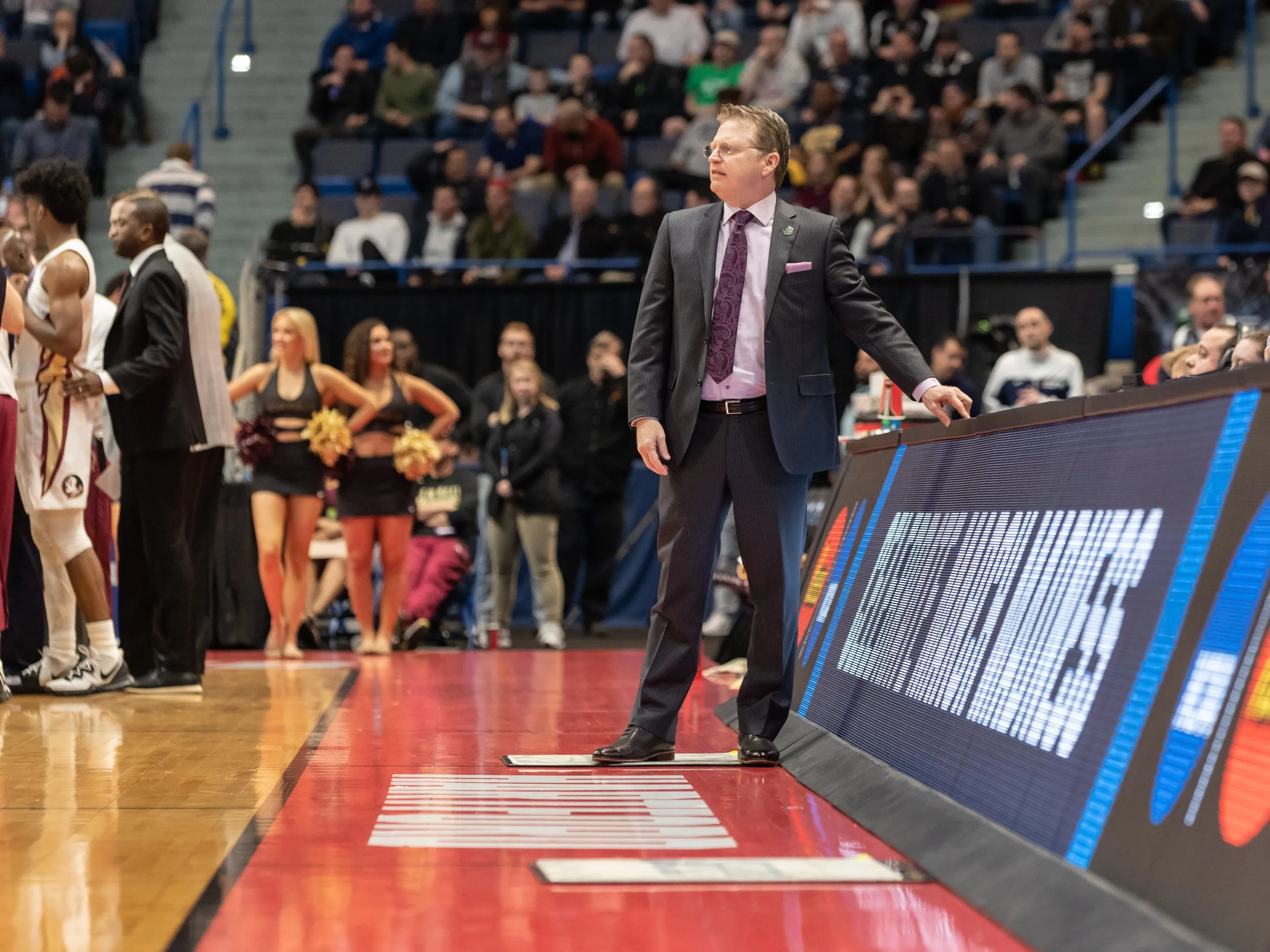 A basketball coach standing on the sideline during a game, with players and cheerleaders visible in the background.