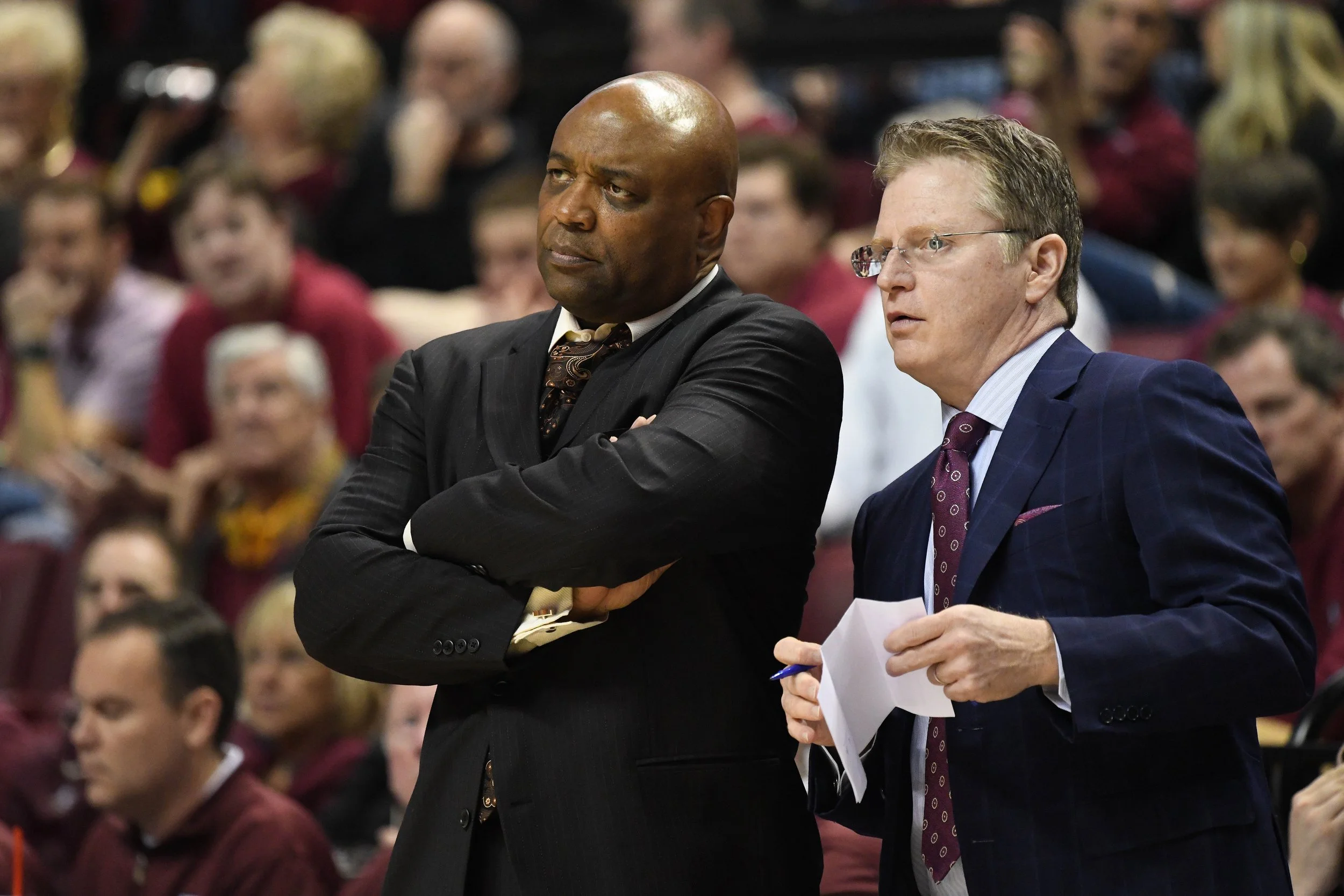 Two men in suits standing and talking in front of a crowd, one with arms crossed and the other holding paper and pen, in a large indoor setting.