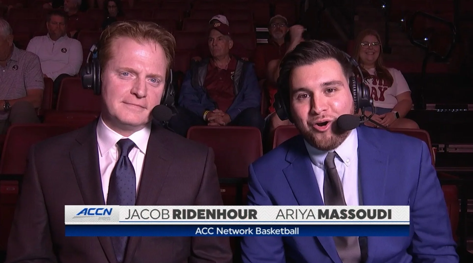 Two male sports broadcasters in suits with headsets and microphones, sitting courtside at a basketball game, with a crowd seated behind them. Text overlay shows their names: Jacob Ridenhour and Ariya Massoudi, and mentions the ACC Network Basketball.