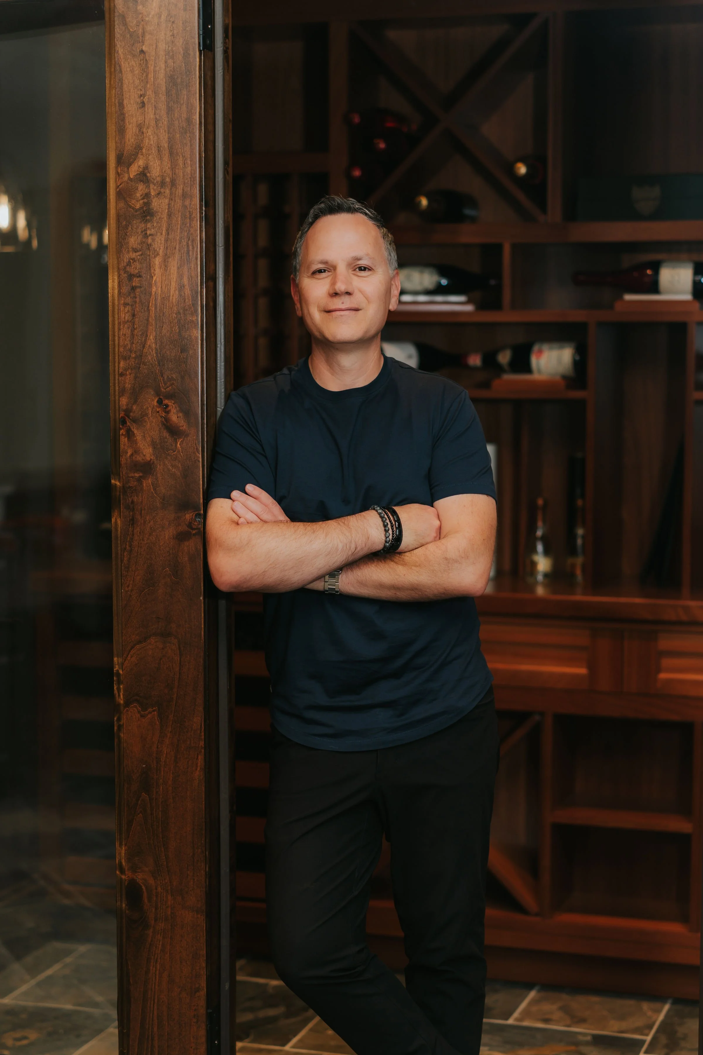 A man standing with arms crossed, leaning against a wooden wall in a wine cellar.