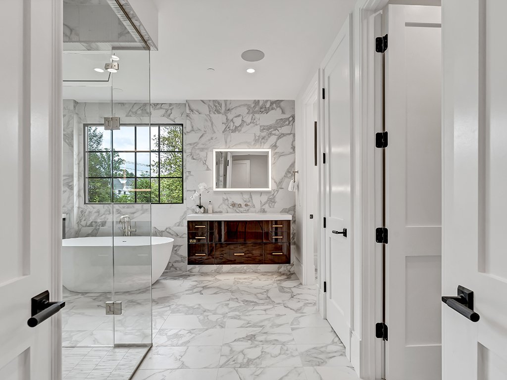 Modern bathroom with marble floors and walls, a freestanding bathtub near a window, a glass-enclosed shower, a wooden vanity with a mirror, and white doors.