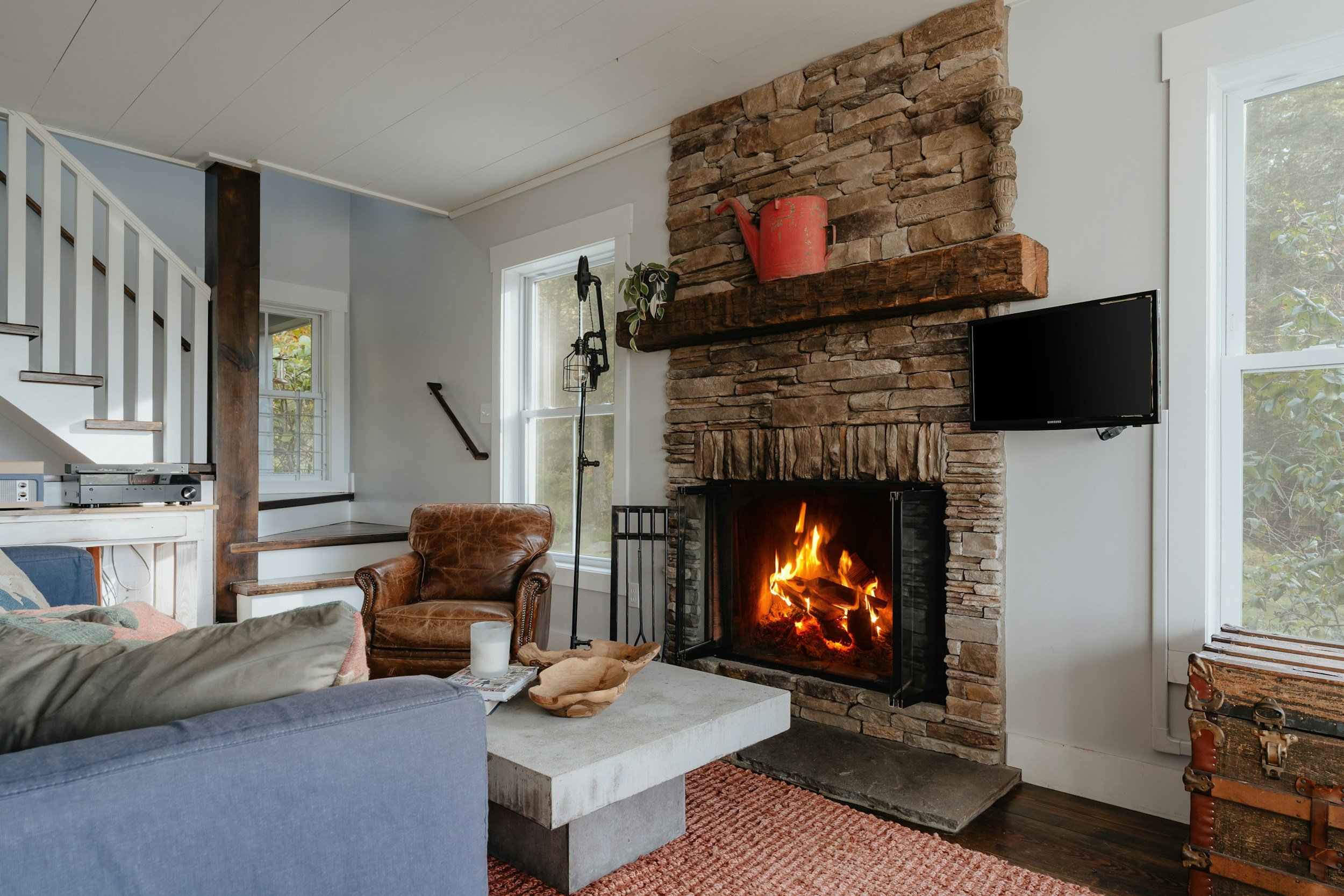 Cozy living room with a glowing fireplace, a flat-screen TV on the wall, a leather armchair, a concrete coffee table with decorative bowls, and large windows with views of trees outside.