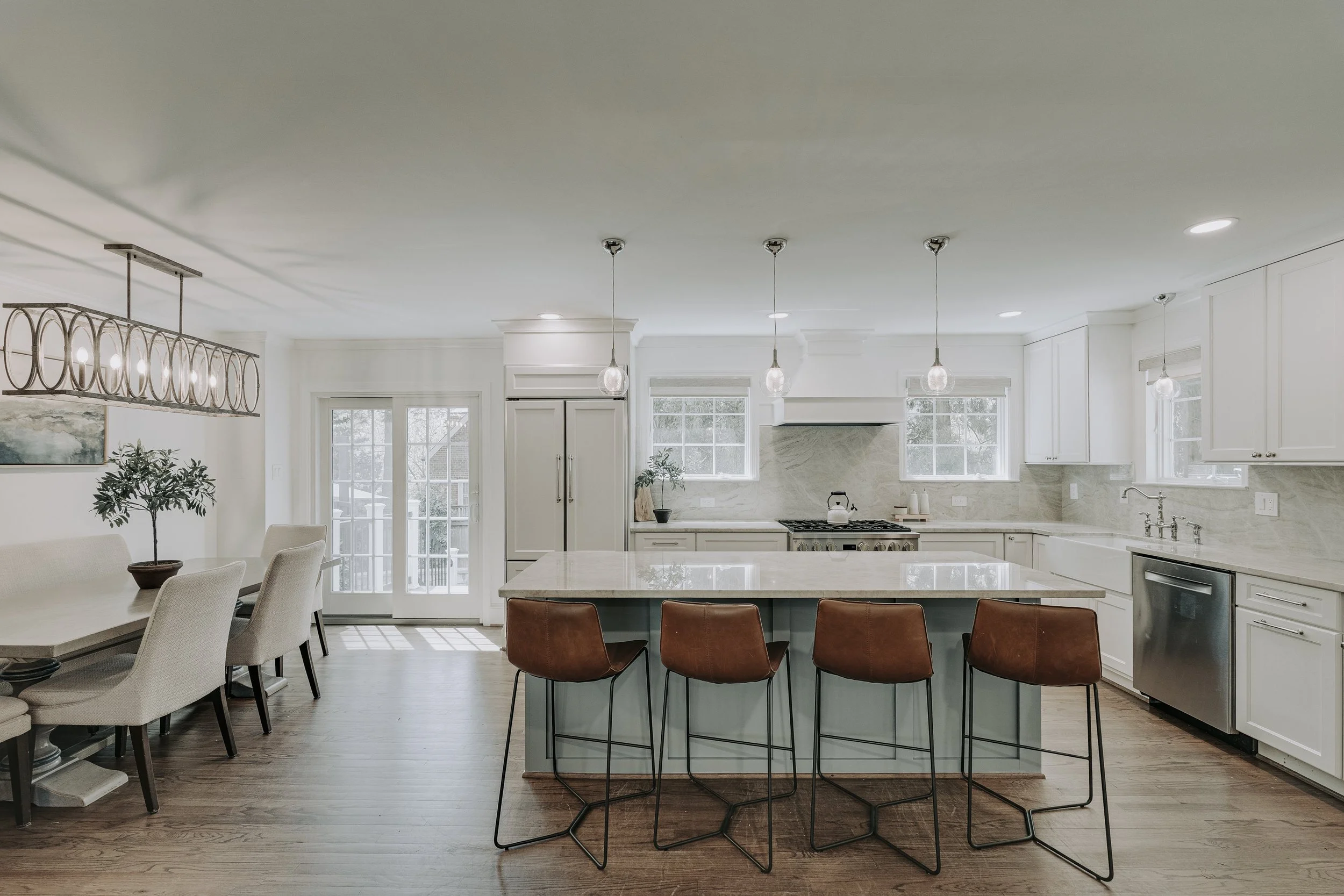 Modern kitchen with white cabinets, marble countertops, and a large central island with four brown bar stools. Open dining area with a table and cream-colored chairs. Hardwood floors, pendant lights over the island, and a sliding glass door leading outside.