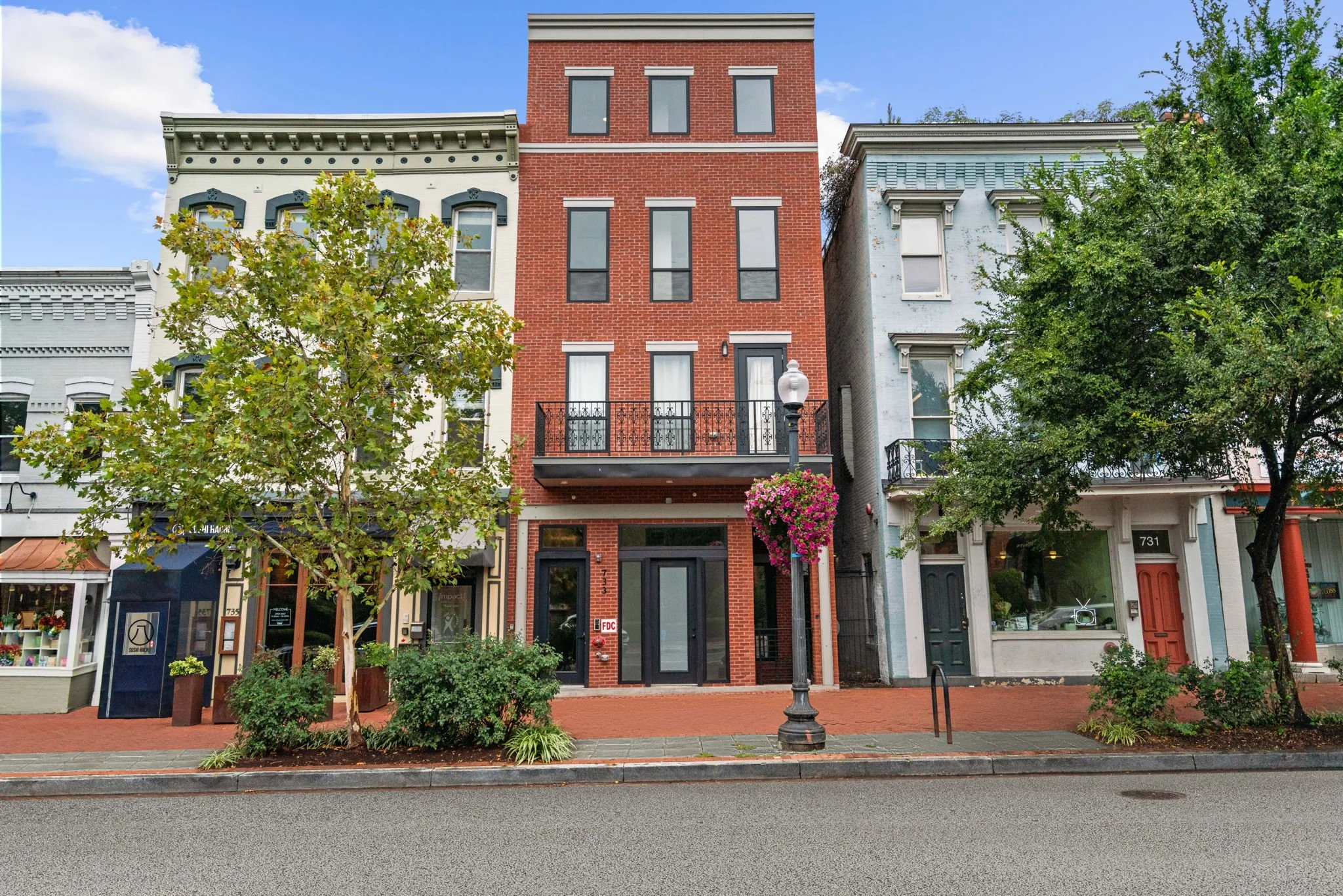 A row of colorful buildings along a street, with trees, sidewalk, and a street lamp in the foreground. The buildings vary in architectural style and color, including green, red brick, and light blue.