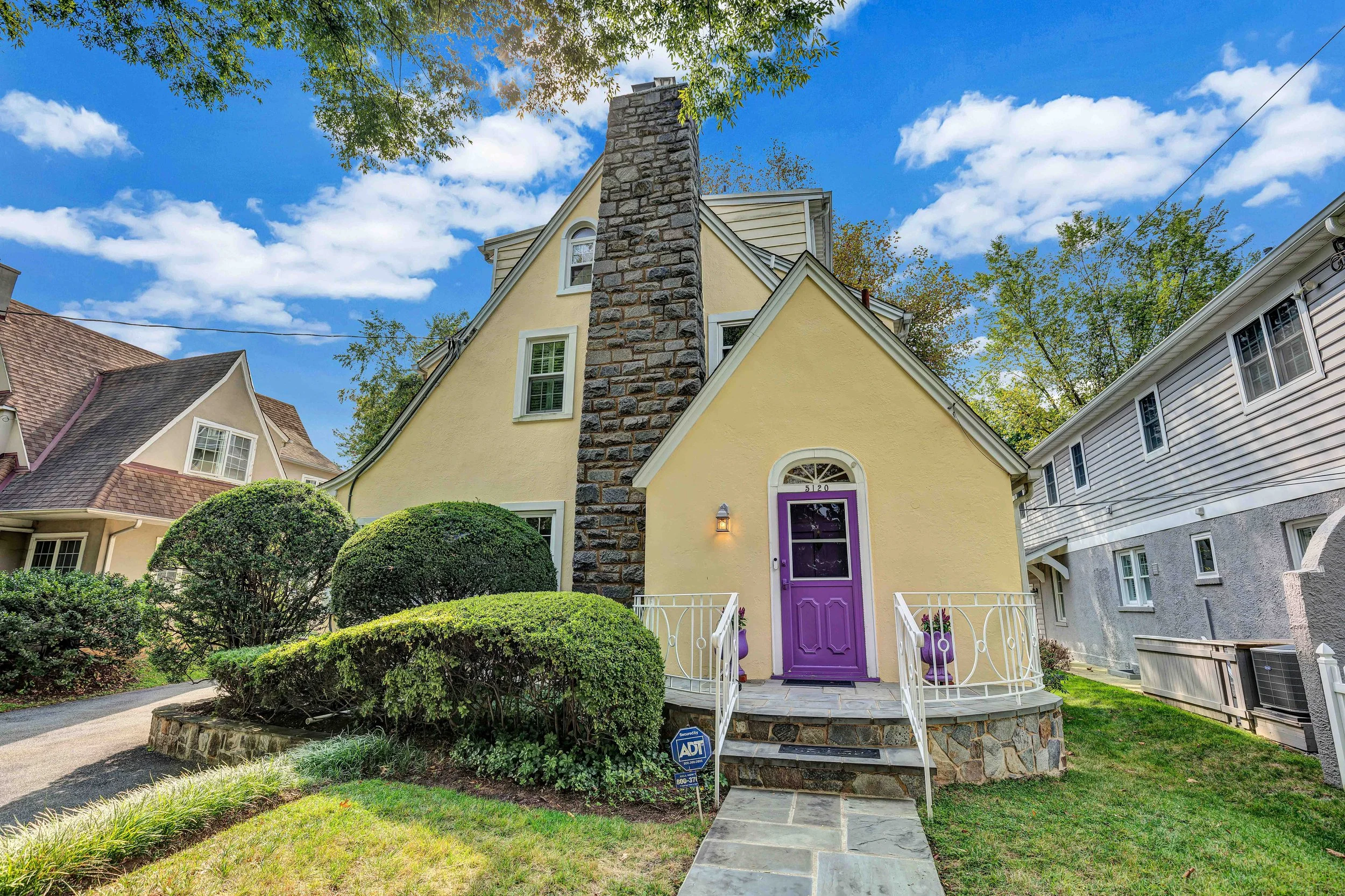 A yellow house with a purple front door, stone chimney, and manicured bushes in a neighborhood on a sunny day.