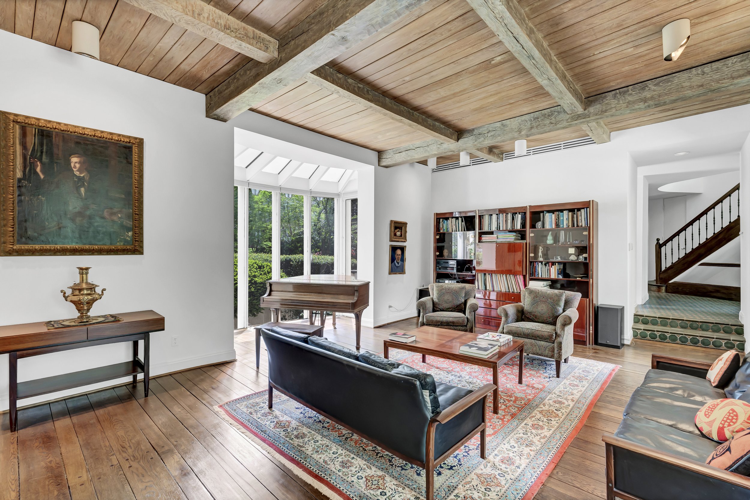 Living room with wooden ceiling, white walls, hardwood floor, and large windows. Contains a grand piano near the window, a sofa, two armchairs, a coffee table with books, a bookshelf with various items, and a portrait painting on the wall.