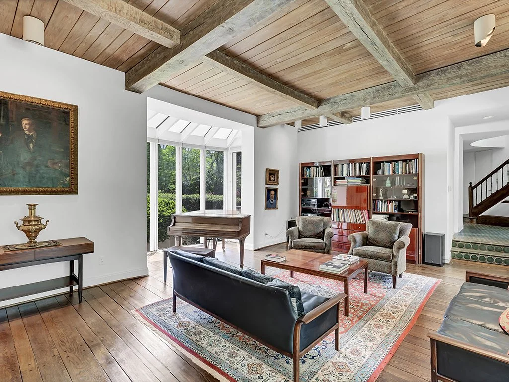 Living room with wooden floors and ceiling beams, featuring a piano near large windows, a black sofa, two armchairs, a coffee table, a wooden bookshelf, and framed artwork on the walls.
