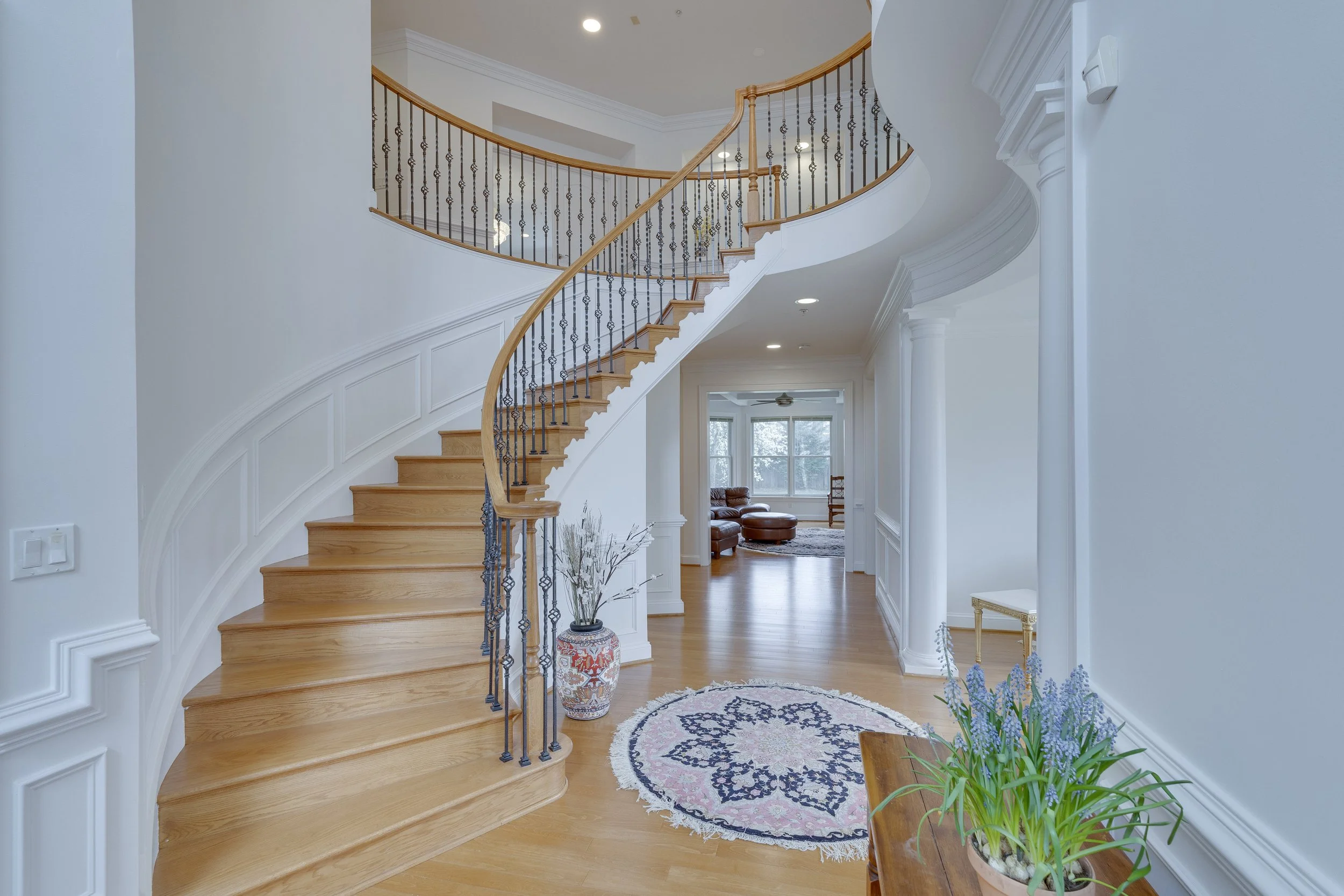 Interior view of a home with a curved staircase, hardwood floors, and white walls, decorated with plants and a circular rug.