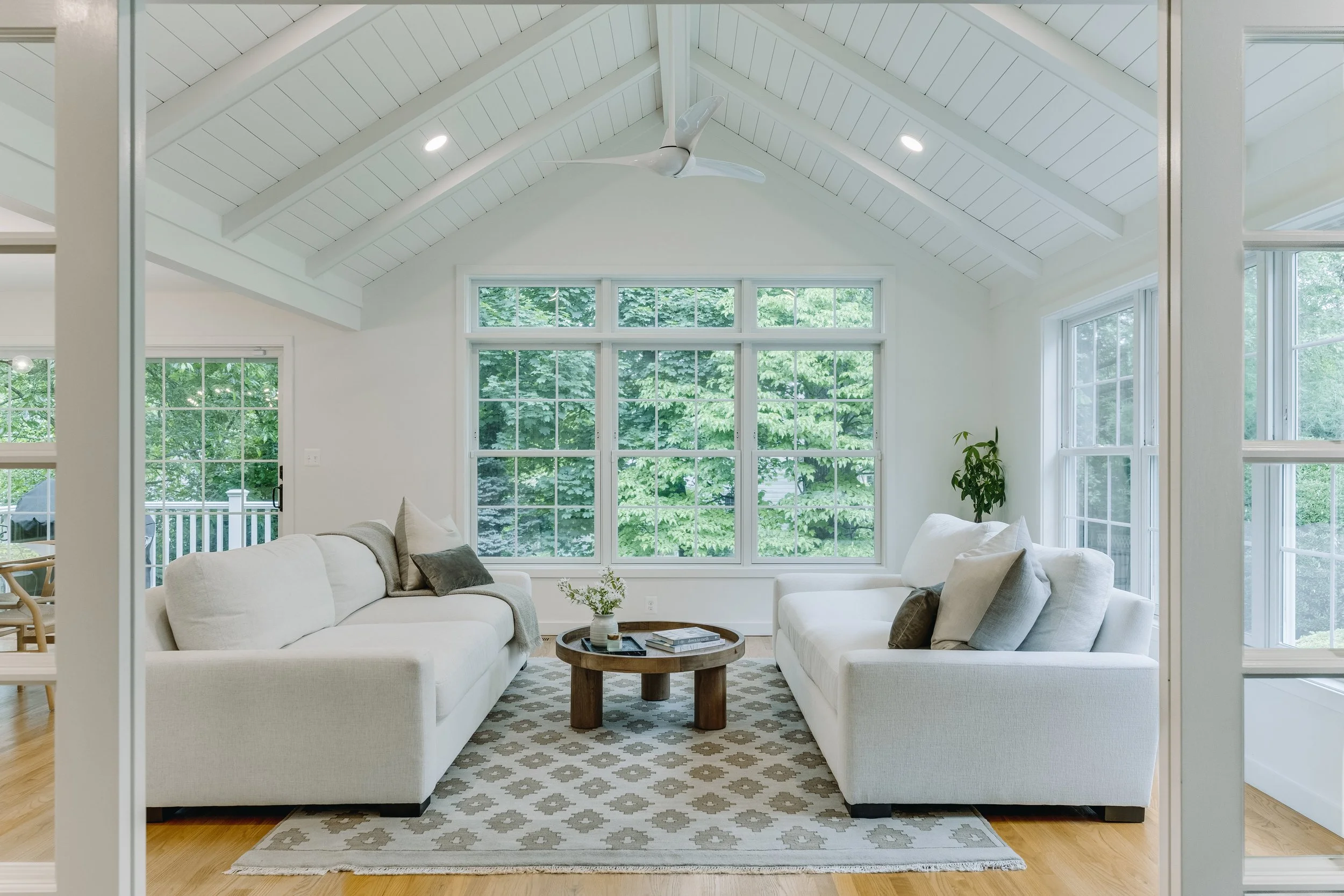 Bright living room with large window, white couches, a wooden coffee table, and a patterned rug.