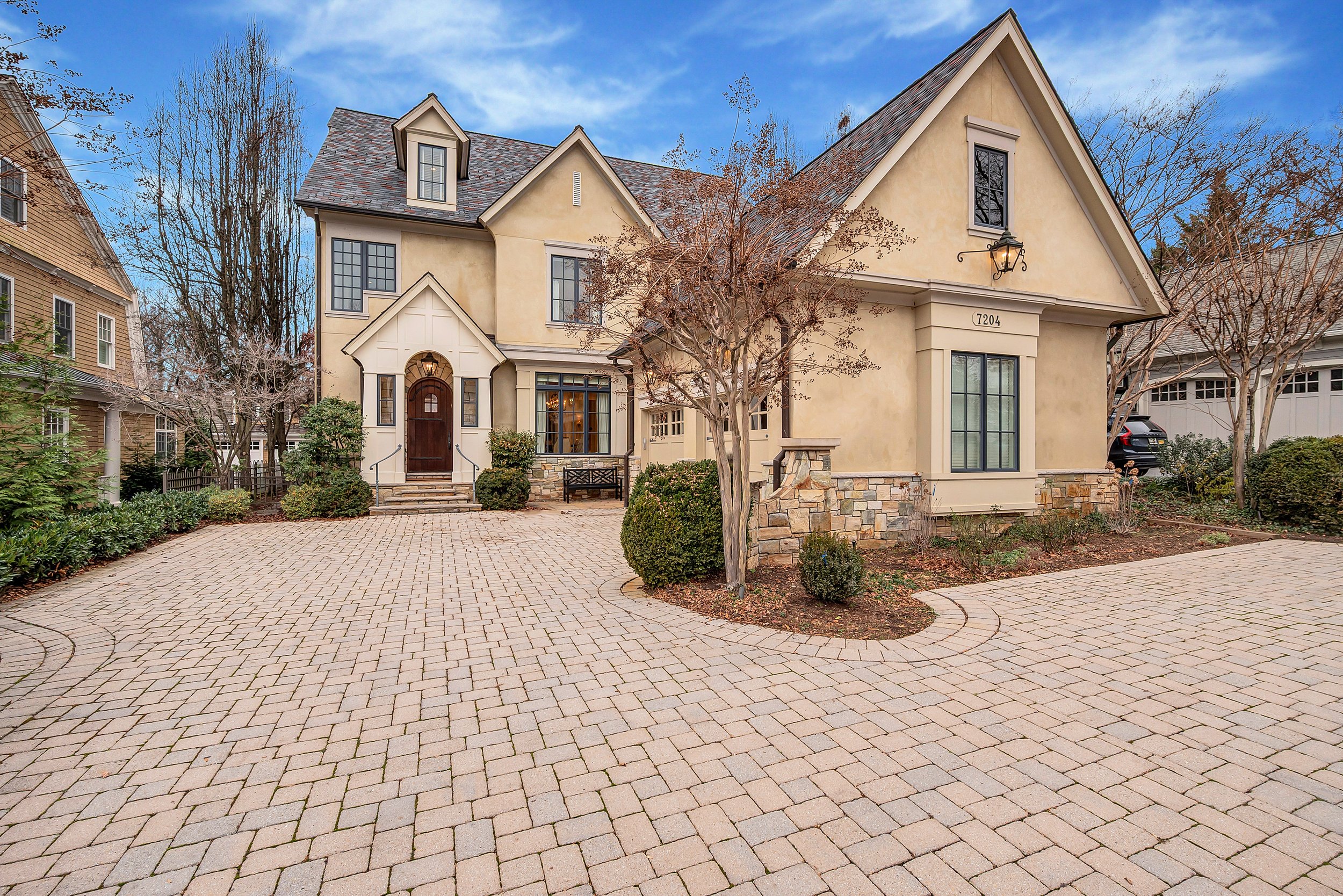 A large, elegant house with a paved driveway, leafless trees, bushes, and a clear blue sky.