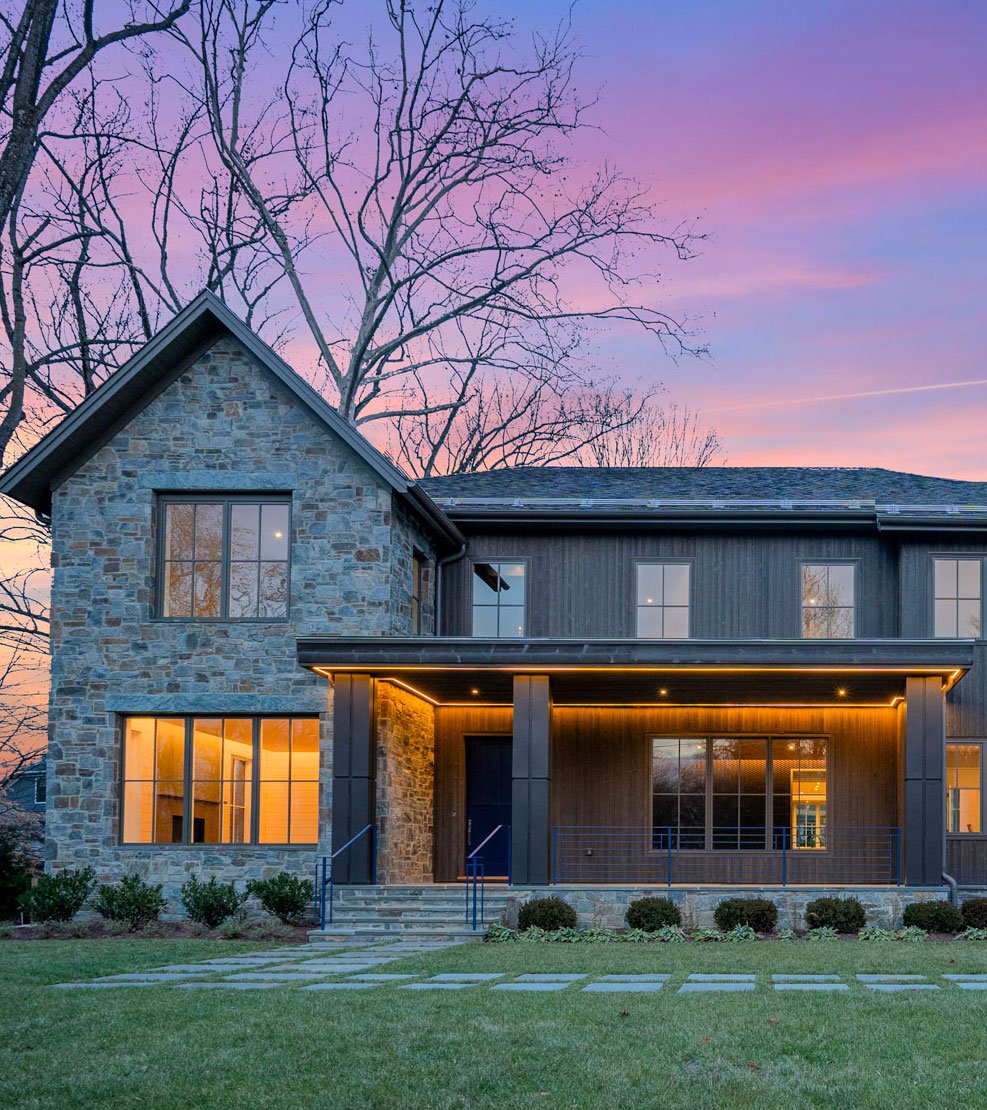 Modern house with stone and dark wood exterior, large windows, and a front porch, set against a sunset sky with pink and purple hues, and trees without leaves in the background.