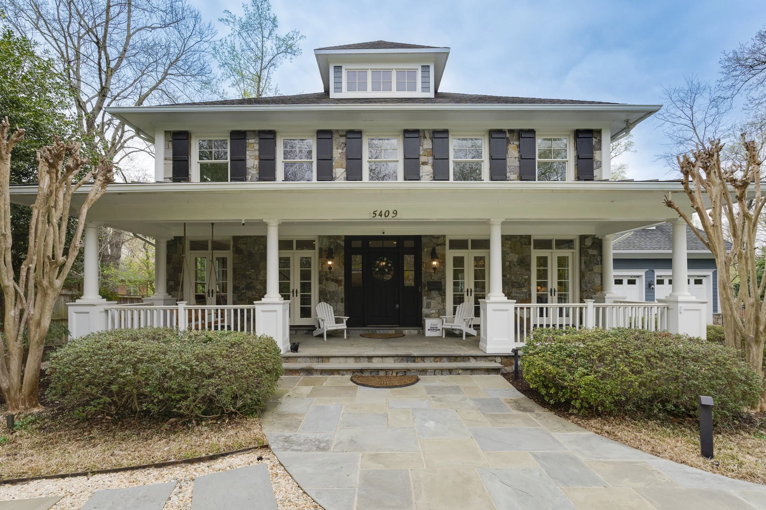 Front view of a large two-story house with stone and white exterior, black shutters, a wide front porch with white railings, white Adirondack chairs, and a stone pathway leading to the entrance. There are bushy shrubs and leafless trees on either side.