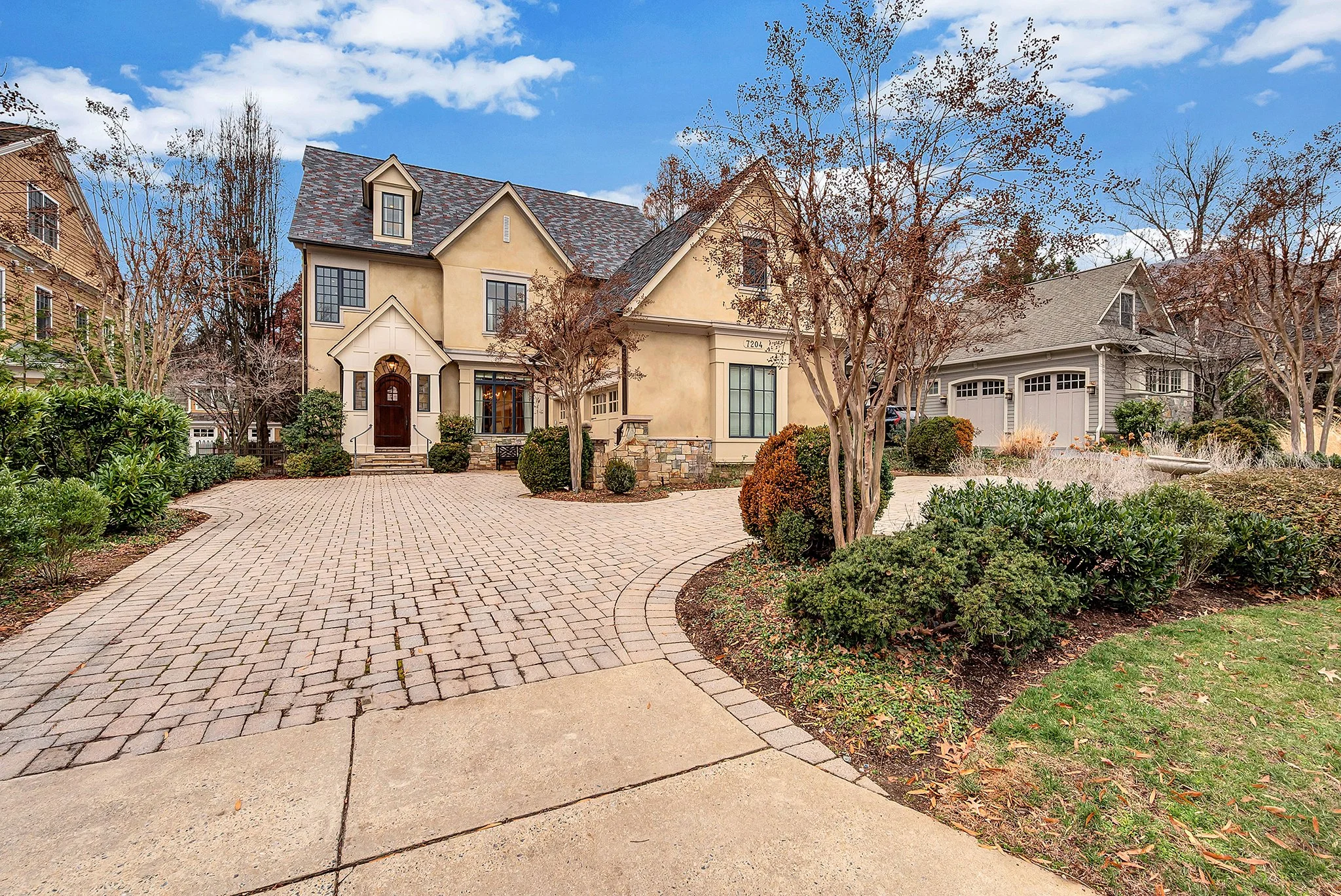A large, elegant house with a paved driveway, surrounded by autumn trees and shrubs, under a partly cloudy sky.