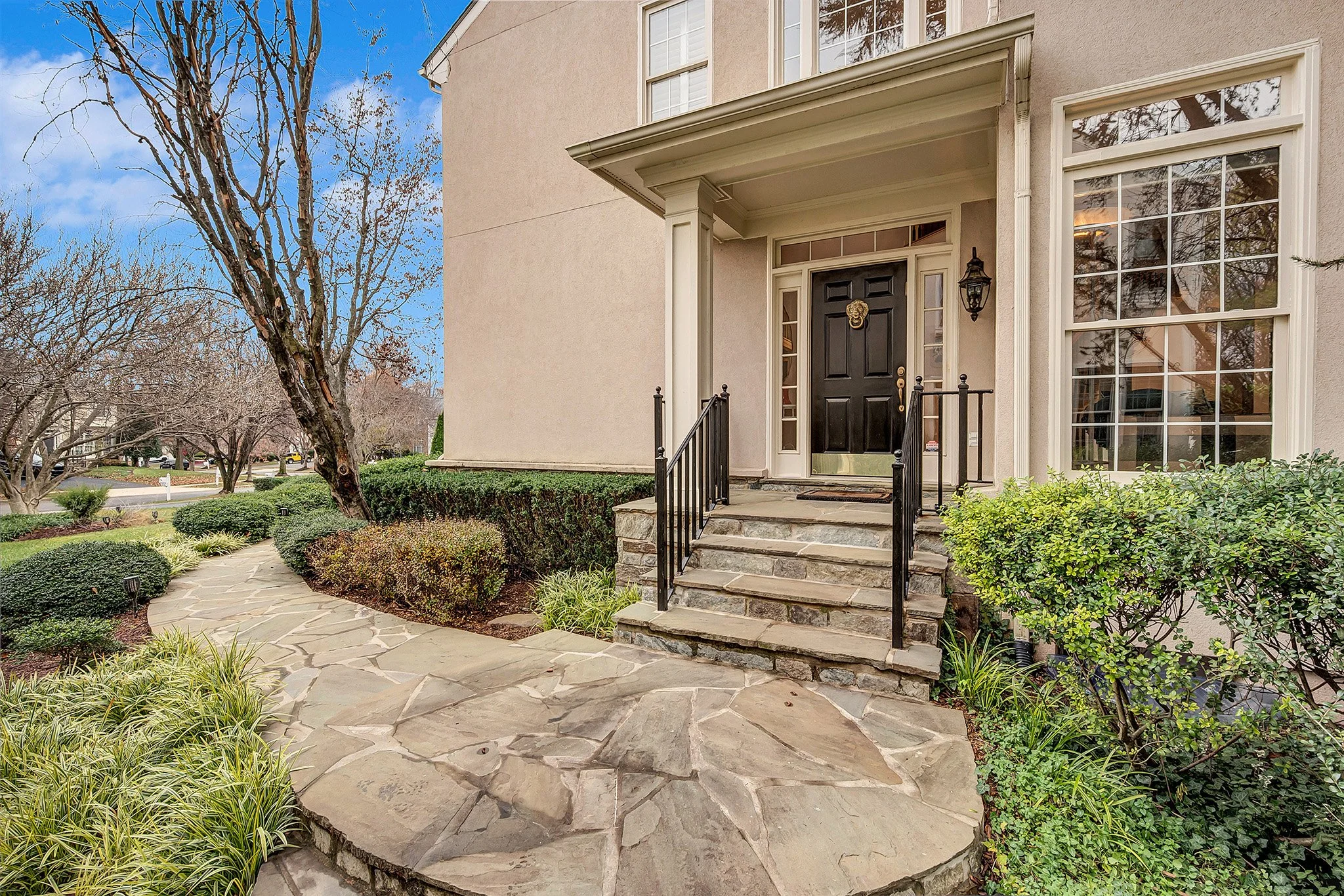 Front entrance of a house with a black door, stone steps, a small porch, and a landscaped yard with bushes, trees, and a stone walkway.