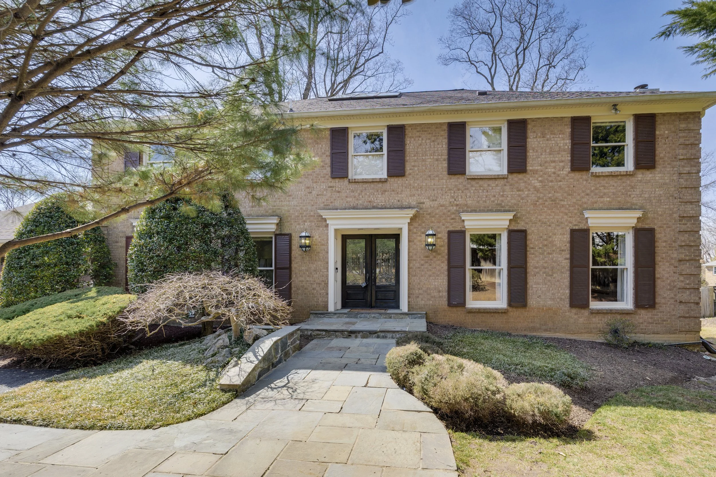 Front view of a two-story brick house with a black front door, brown shutters on five windows, and a stone pathway leading to the entrance, surrounded by well-maintained bushes and trees.