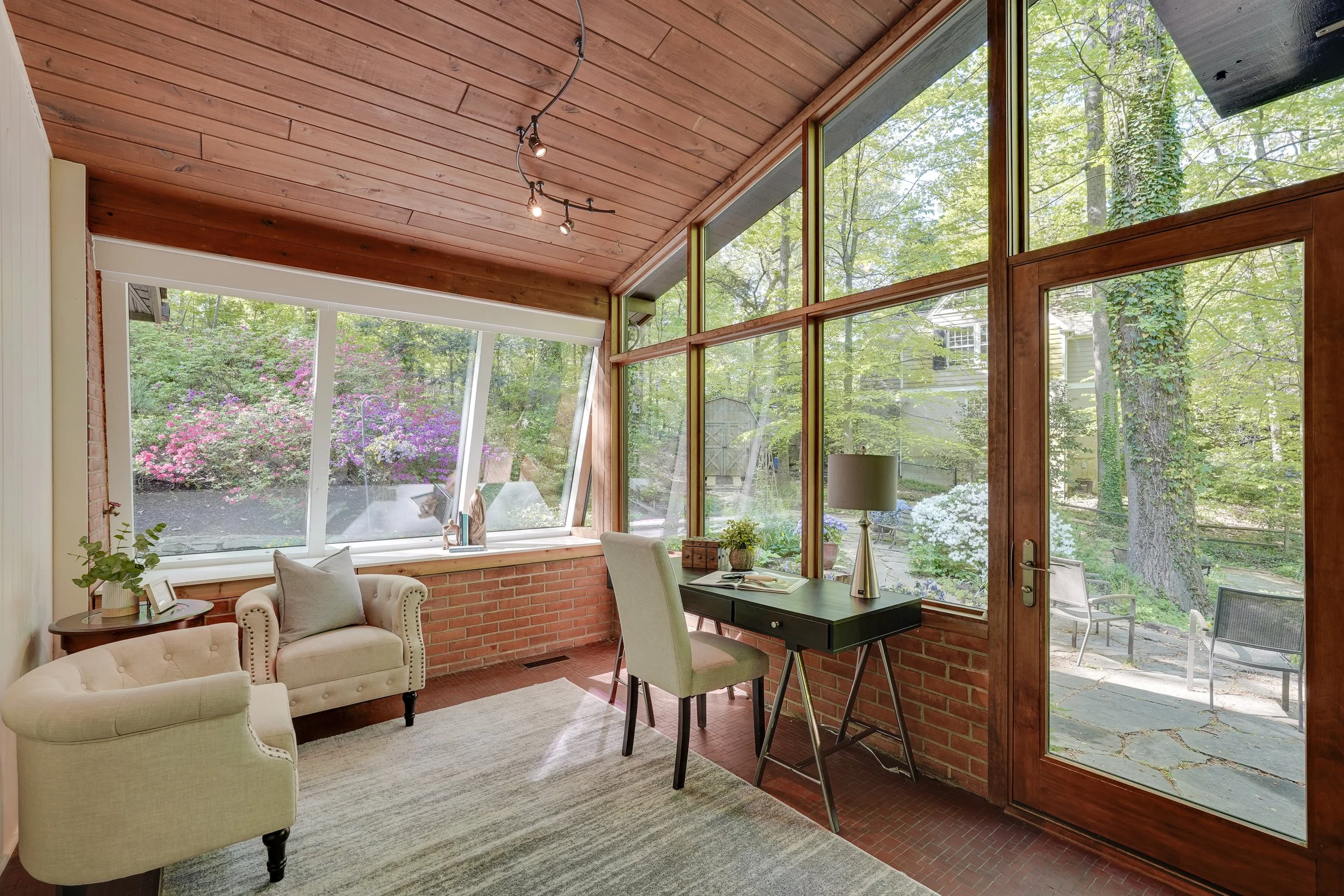 Sunroom with large windows showing a view of a garden with flowering bushes and trees, furnished with beige chairs, a small black desk, and a standing lamp.