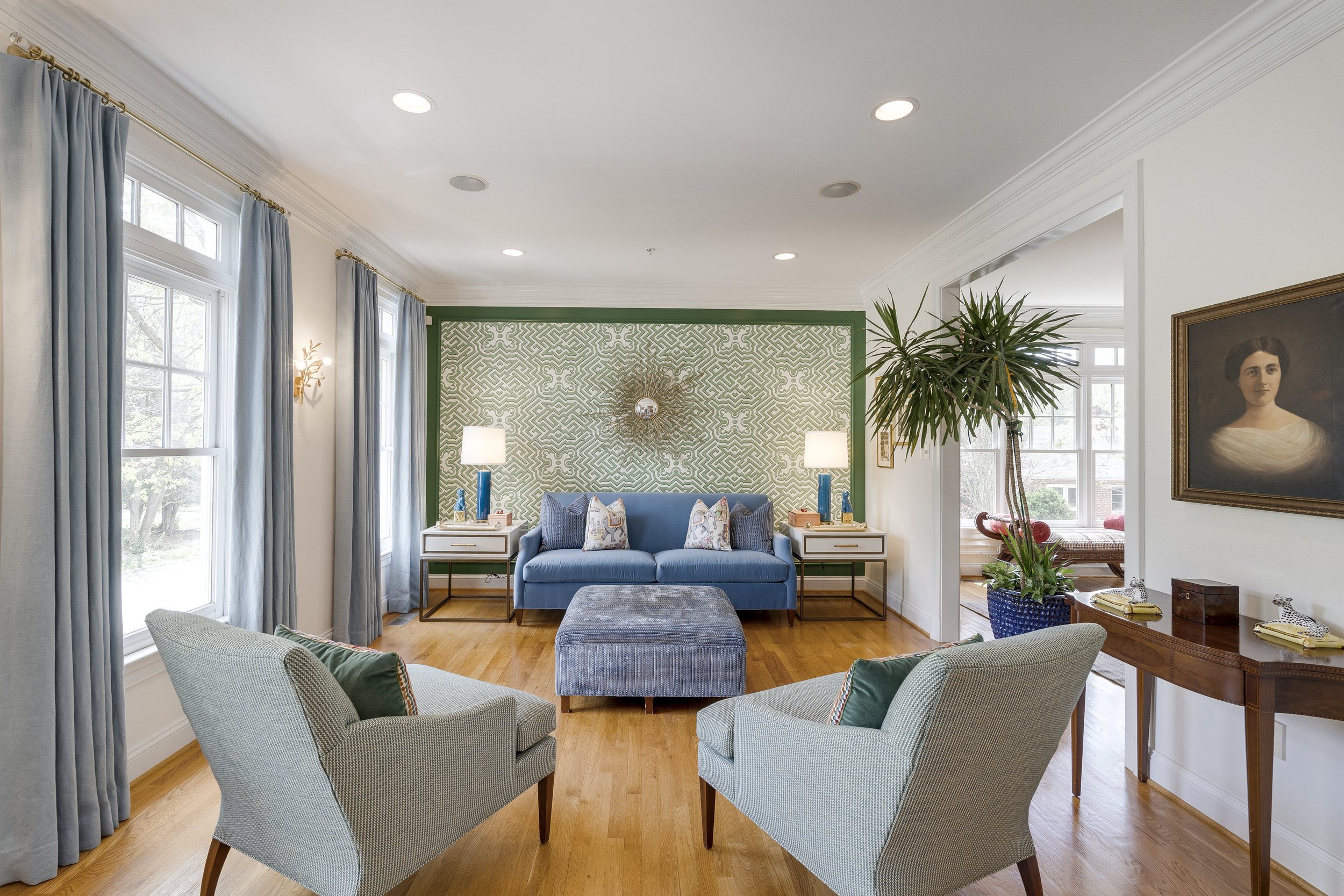 Living room with blue sofa, two armchairs, and two side tables with lamps, large windows with light blue curtains, green patterned accent wall, and wooden floor.