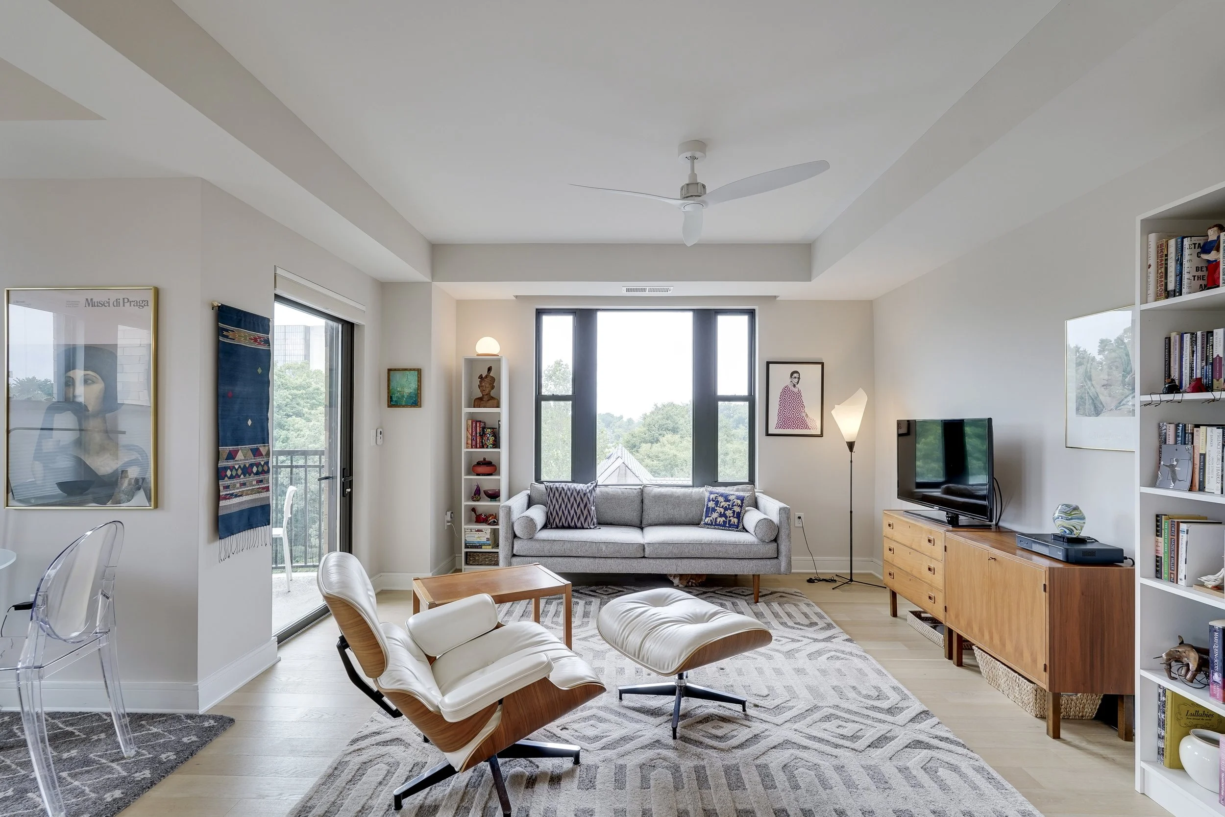 Bright living room with large window, modern white and wood furniture, a gray sofa, white chair with a matching ottoman, a wooden TV stand, bookshelves, artwork, and a sliding door leading to a balcony.
