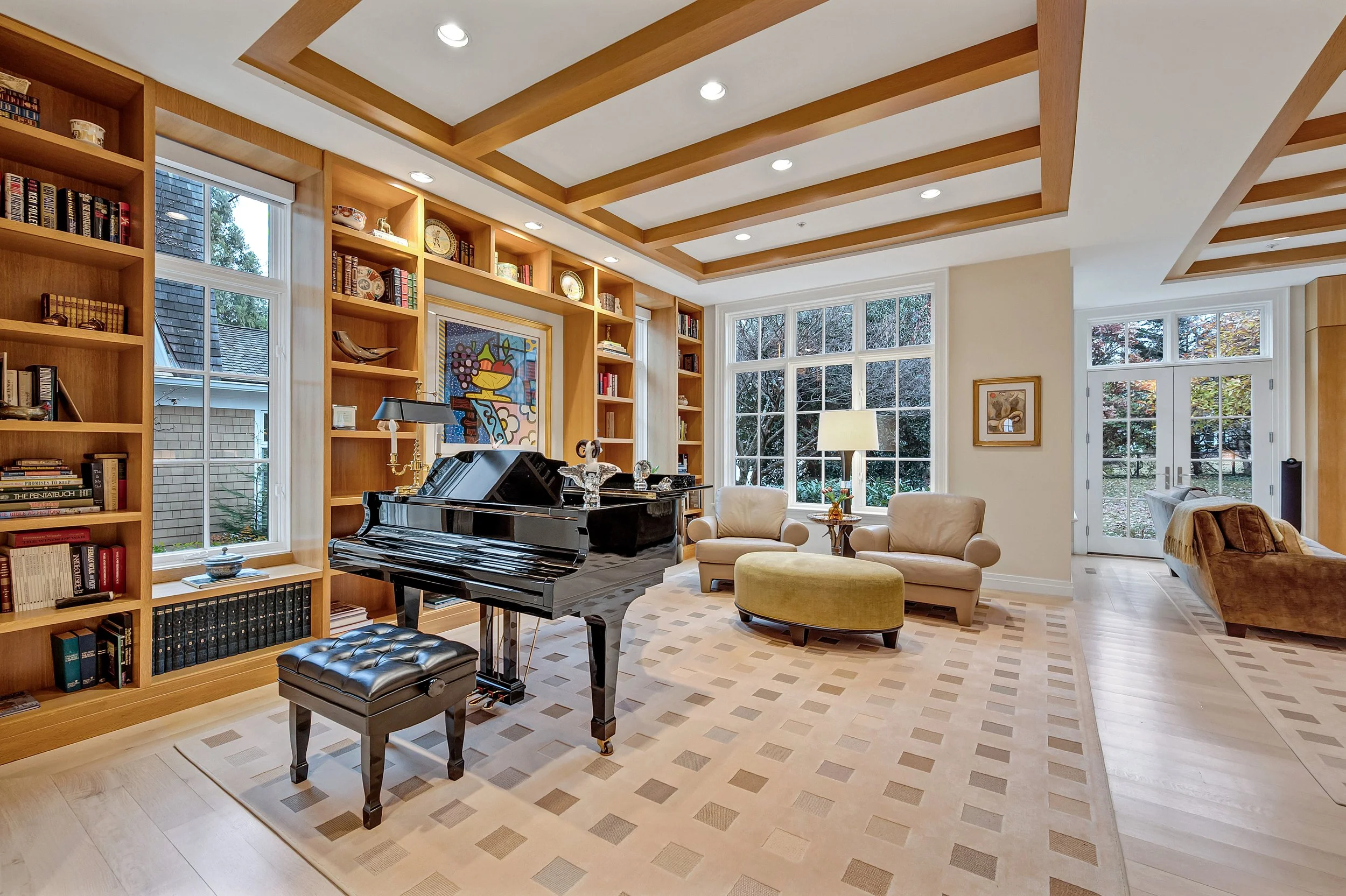Living room with a black grand piano, beige armchairs, a green ottoman, built-in wooden bookshelves, large windows, and a ceiling with wooden beams.