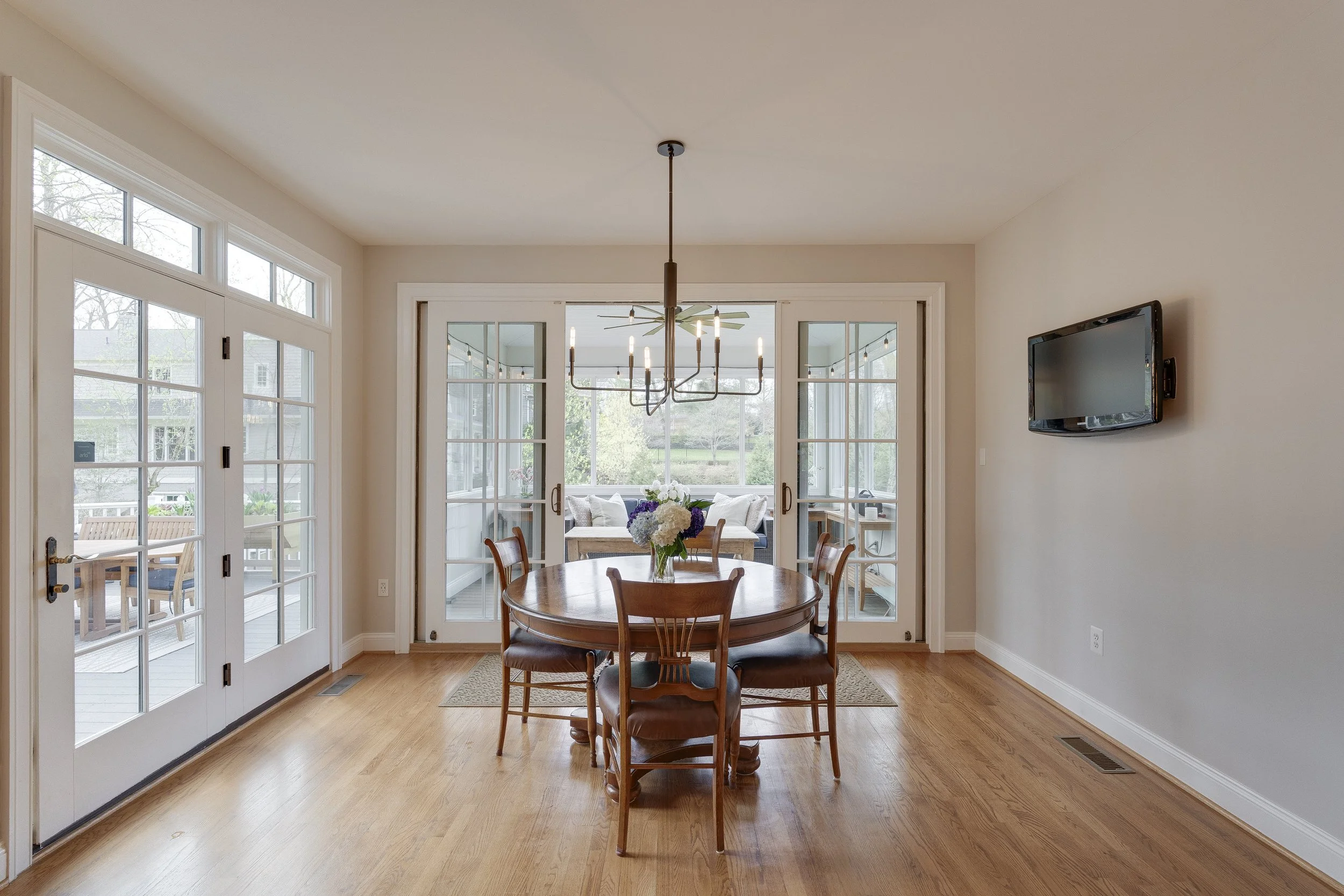Dining room with wooden table and six chairs, a vase with flowers, French doors leading to a patio, and a wall-mounted TV