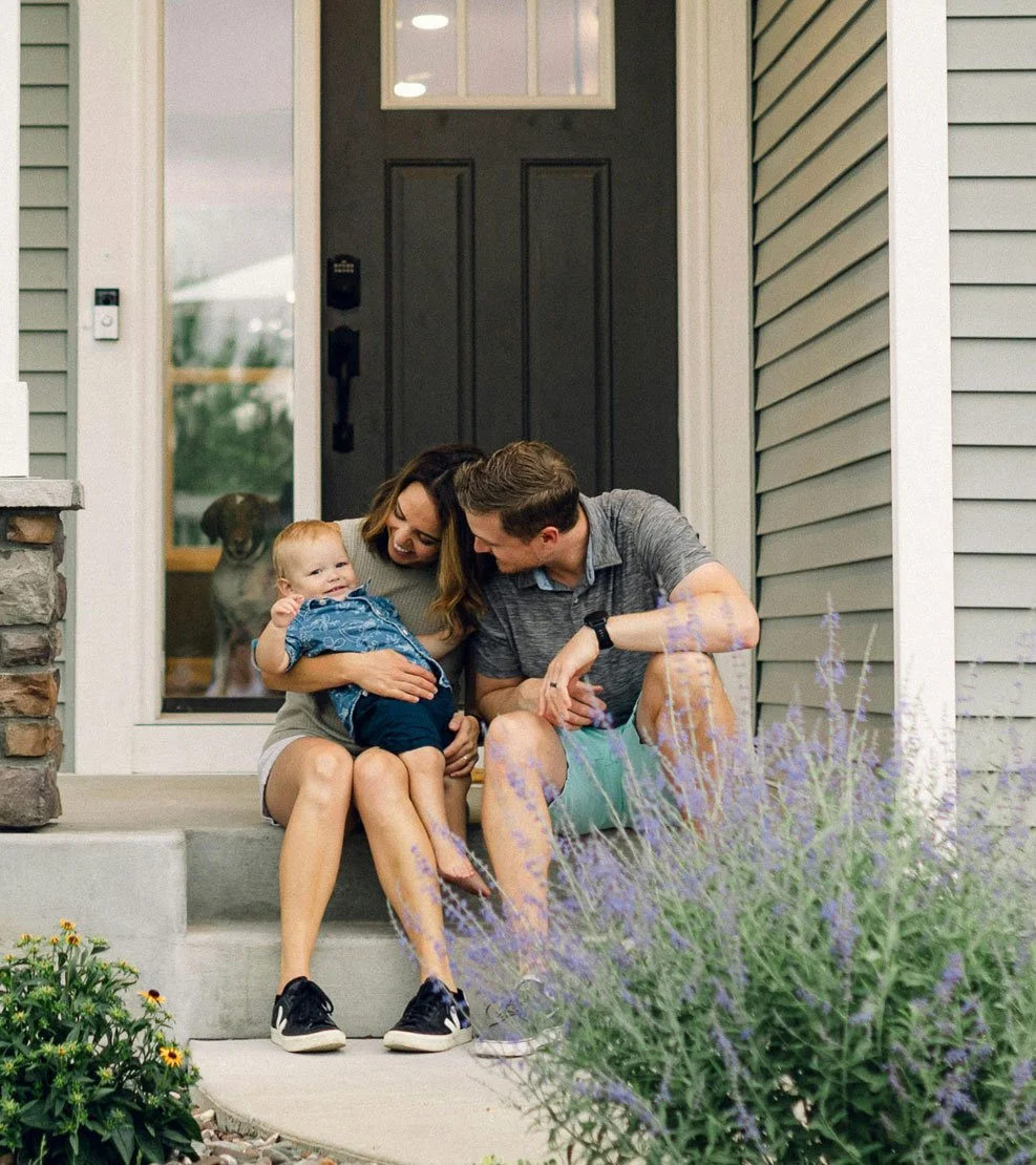 A family of three sitting on the porch steps outside their home, smiling and playing with a baby boy. The mother is holding the baby, and the father is sitting beside them, all enjoying a happy moment.