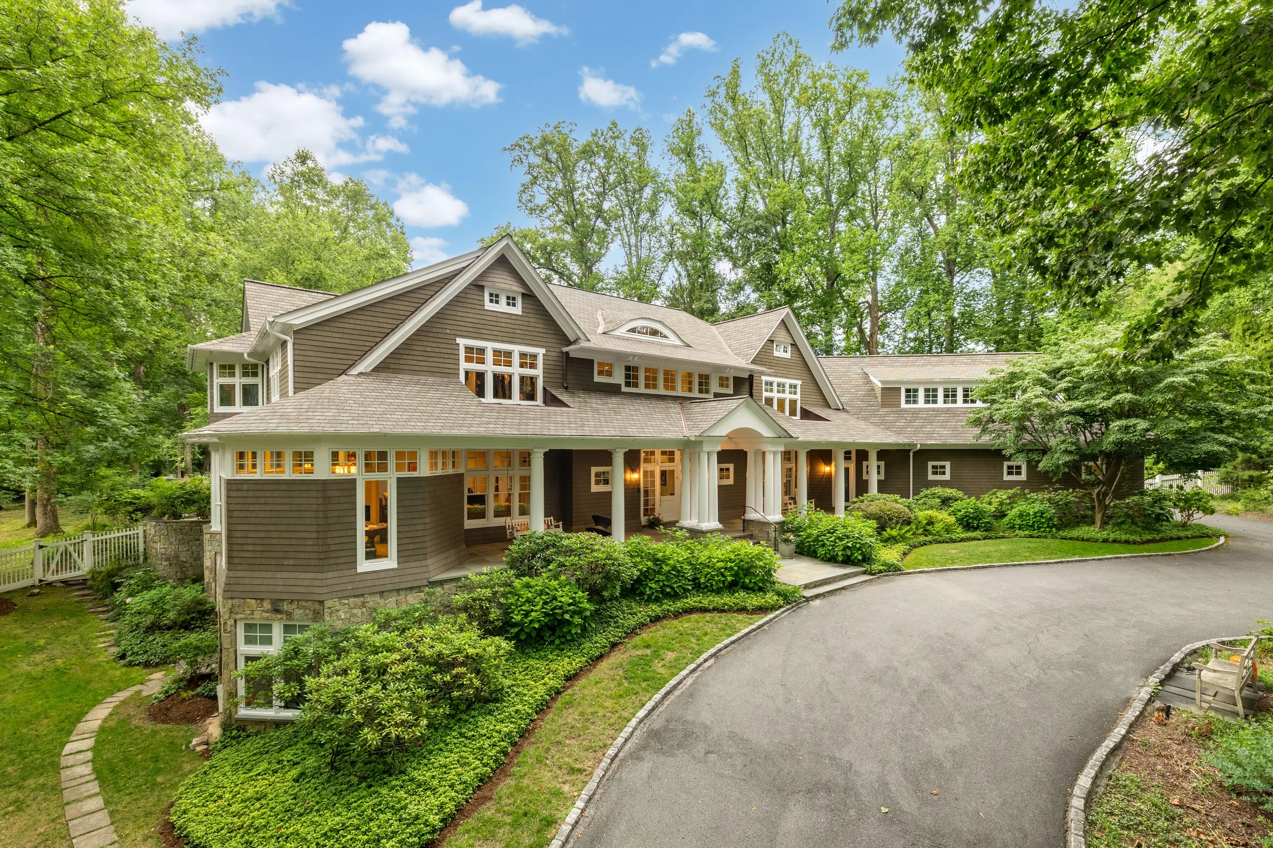 Large, elegant house surrounded by lush green trees and landscaping, with a curved driveway leading to the front entrance, illuminated by exterior lights at dusk.