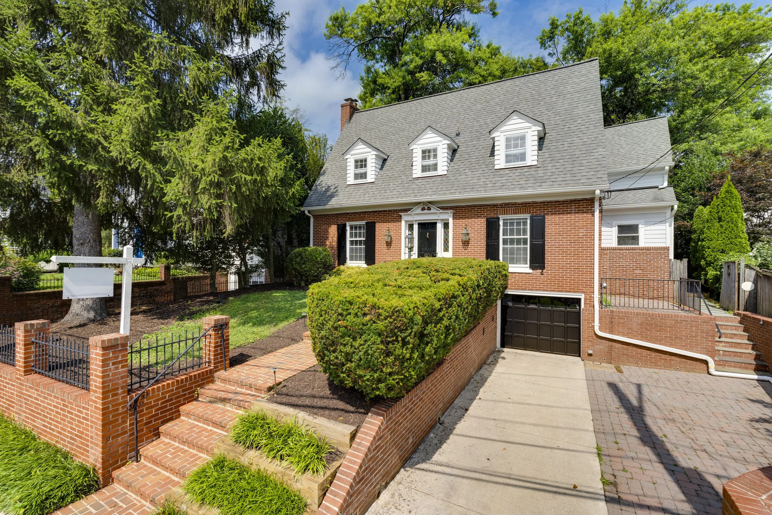 A two-story brick house with black shutters, white trim, and dormer windows, surrounded by greenery and a brick walkway leading to the front door.