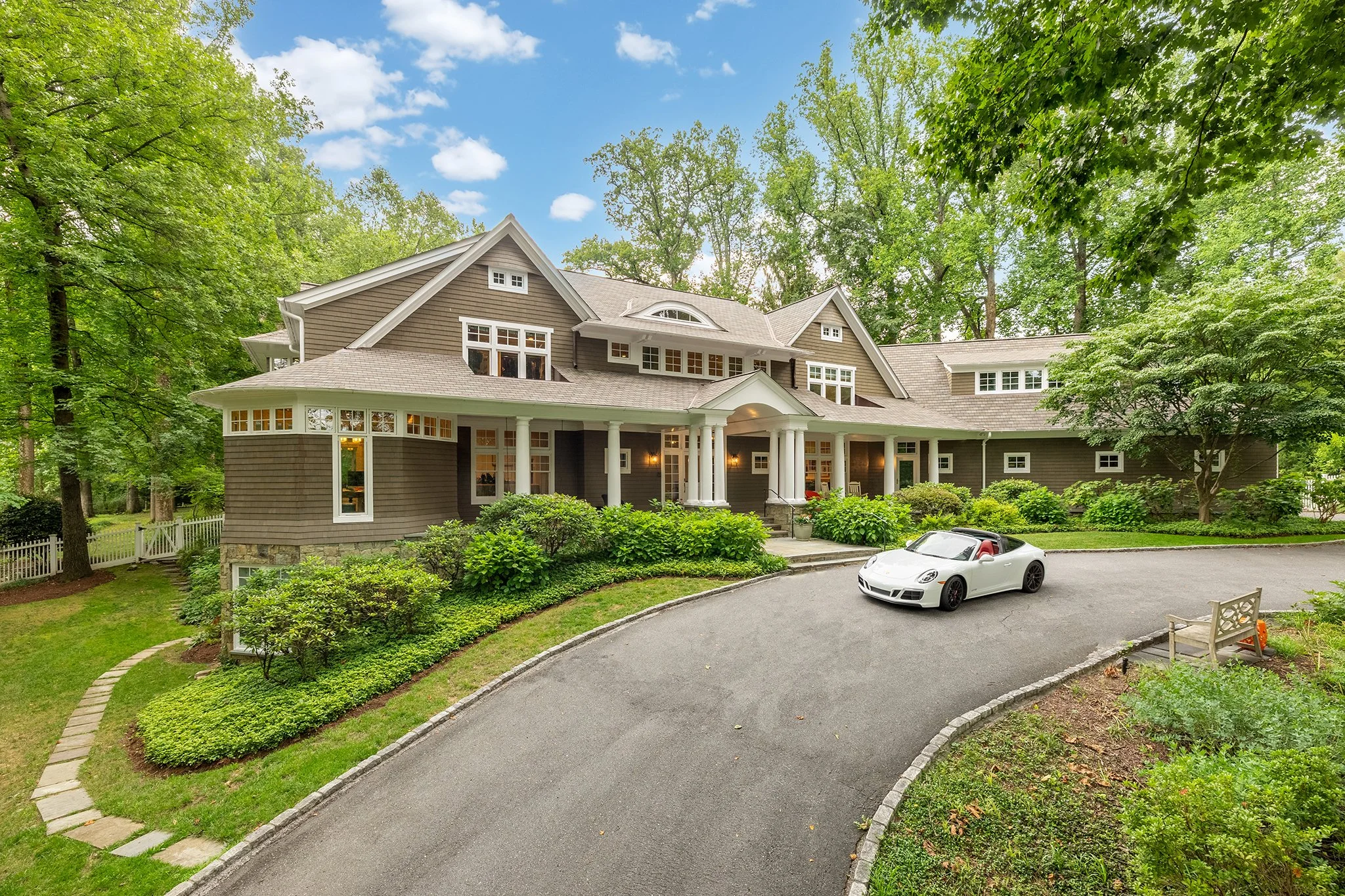 Large house with greenery and a white sports car parked in front, surrounded by tall trees.