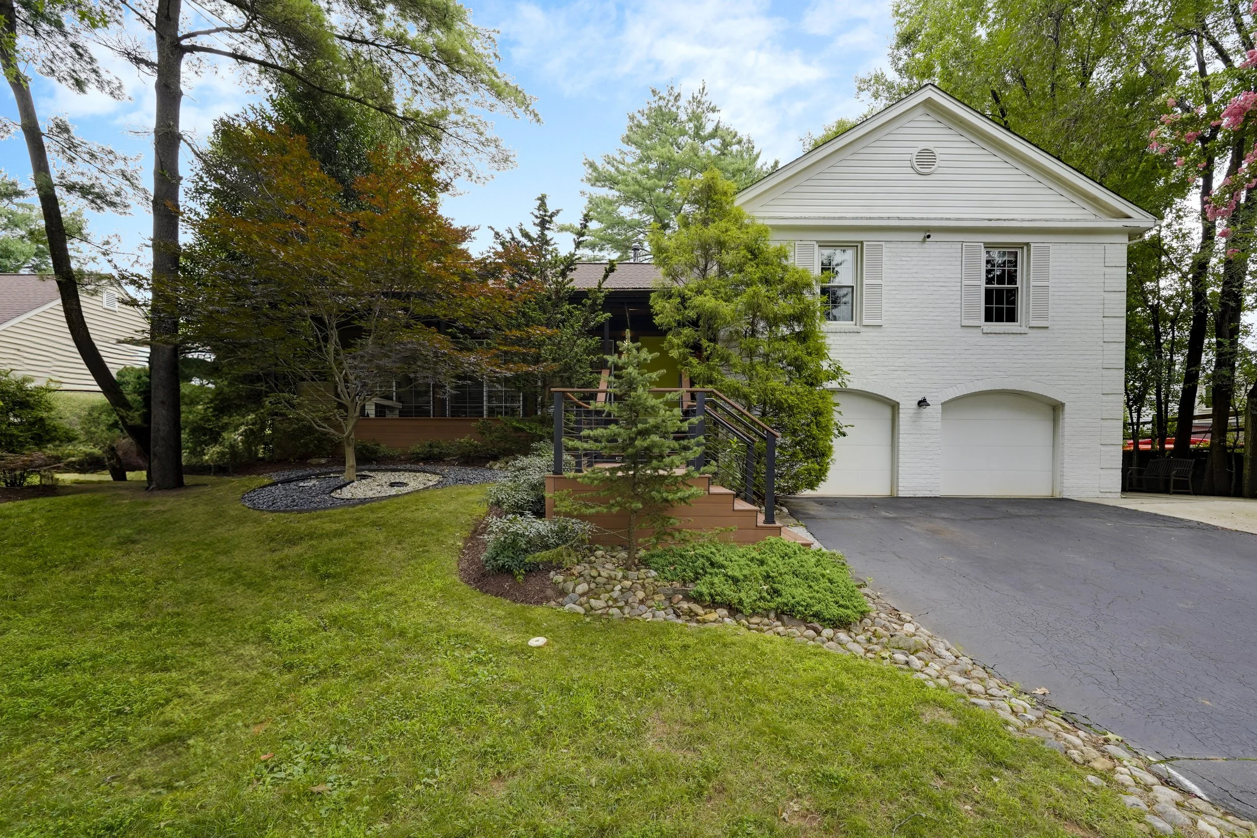 Front yard with green lawn, trees, and shrubs, leading up to a white two-story house with garage doors, a staircase, and a deck surrounded by trees.