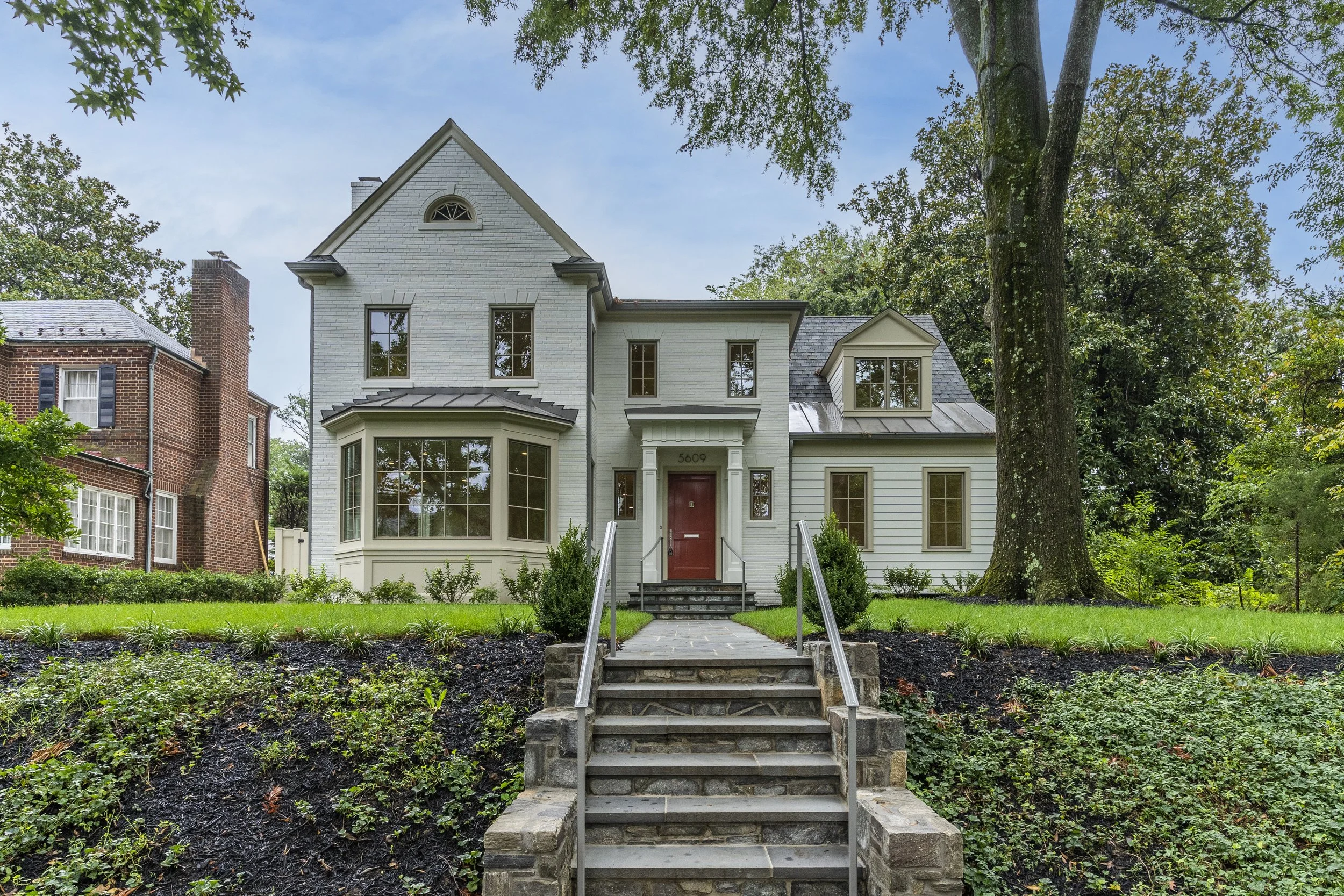 A large white house with multiple floors and various window styles, with stairs leading up to the front door, surrounded by greenery and trees.