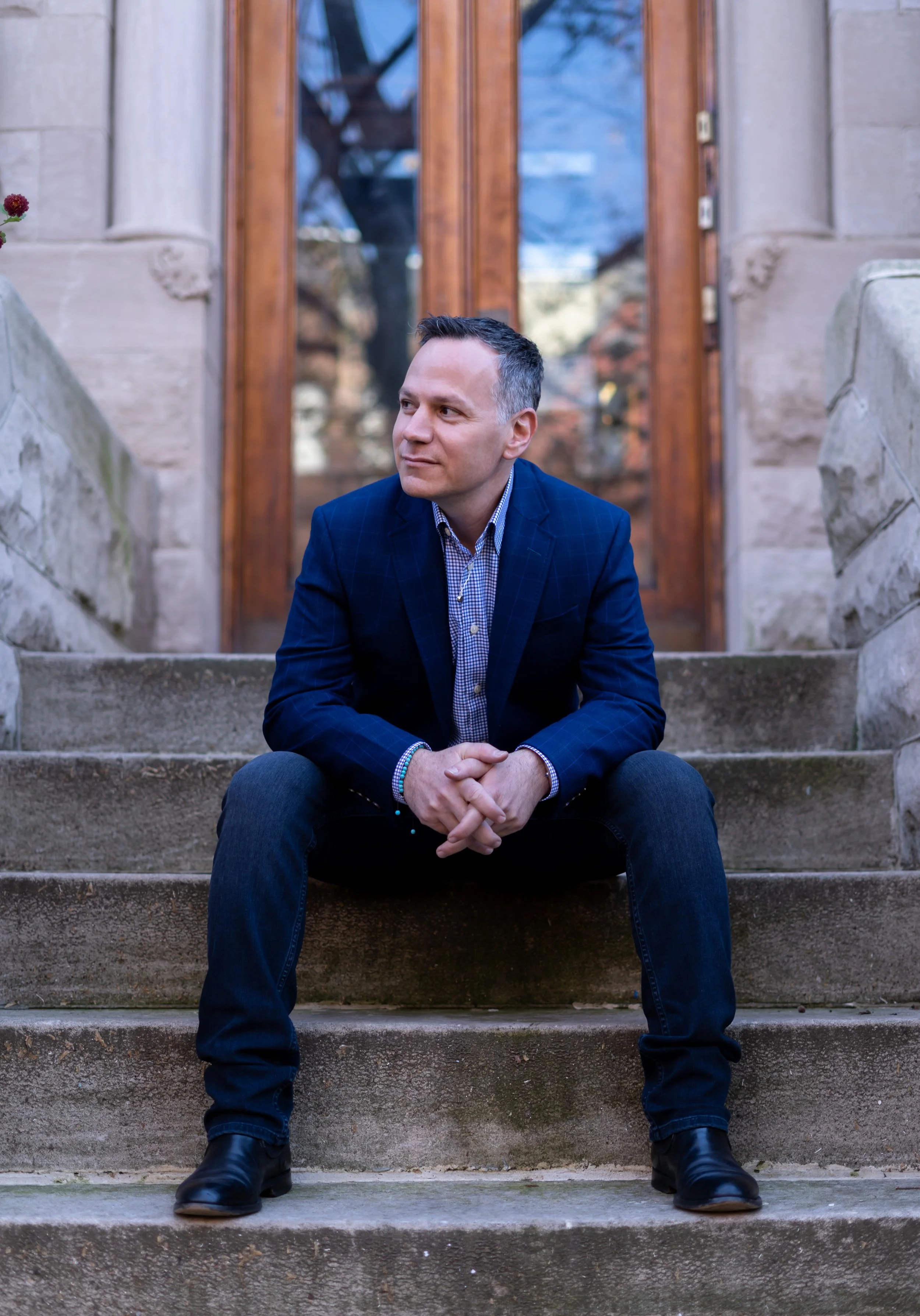 A man in a navy blazer and jeans sitting on stone steps in front of a wooden door.