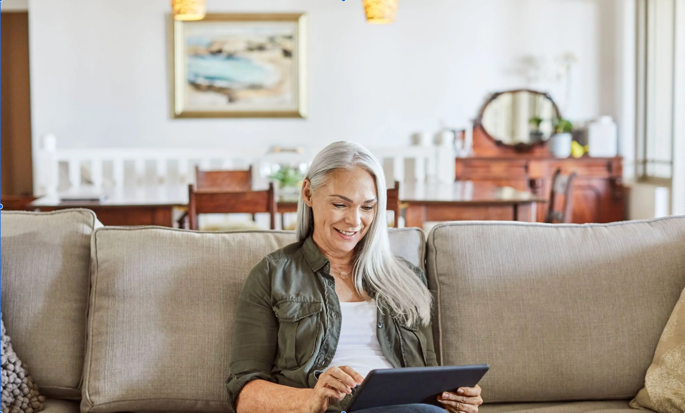 A woman with gray hair sitting on a beige sofa, smiling while looking at a tablet in a bright, cozy living room.