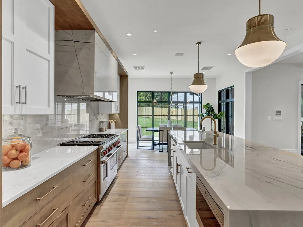 Modern kitchen with white cabinets, marble countertops, a large island with a sink, pendant lights, and a view of a dining area and backyard through large glass doors.