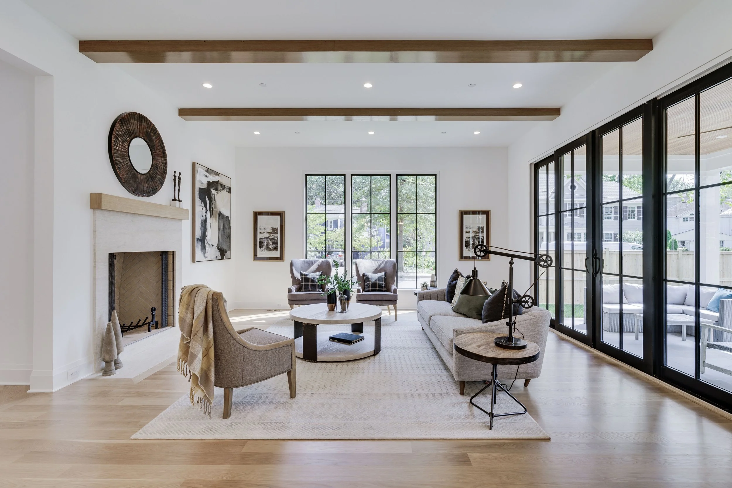 Bright living room with large glass sliding doors, fireplace, modern artwork, beige sofas, armchair, round coffee table, decorative items, and ceiling beams.