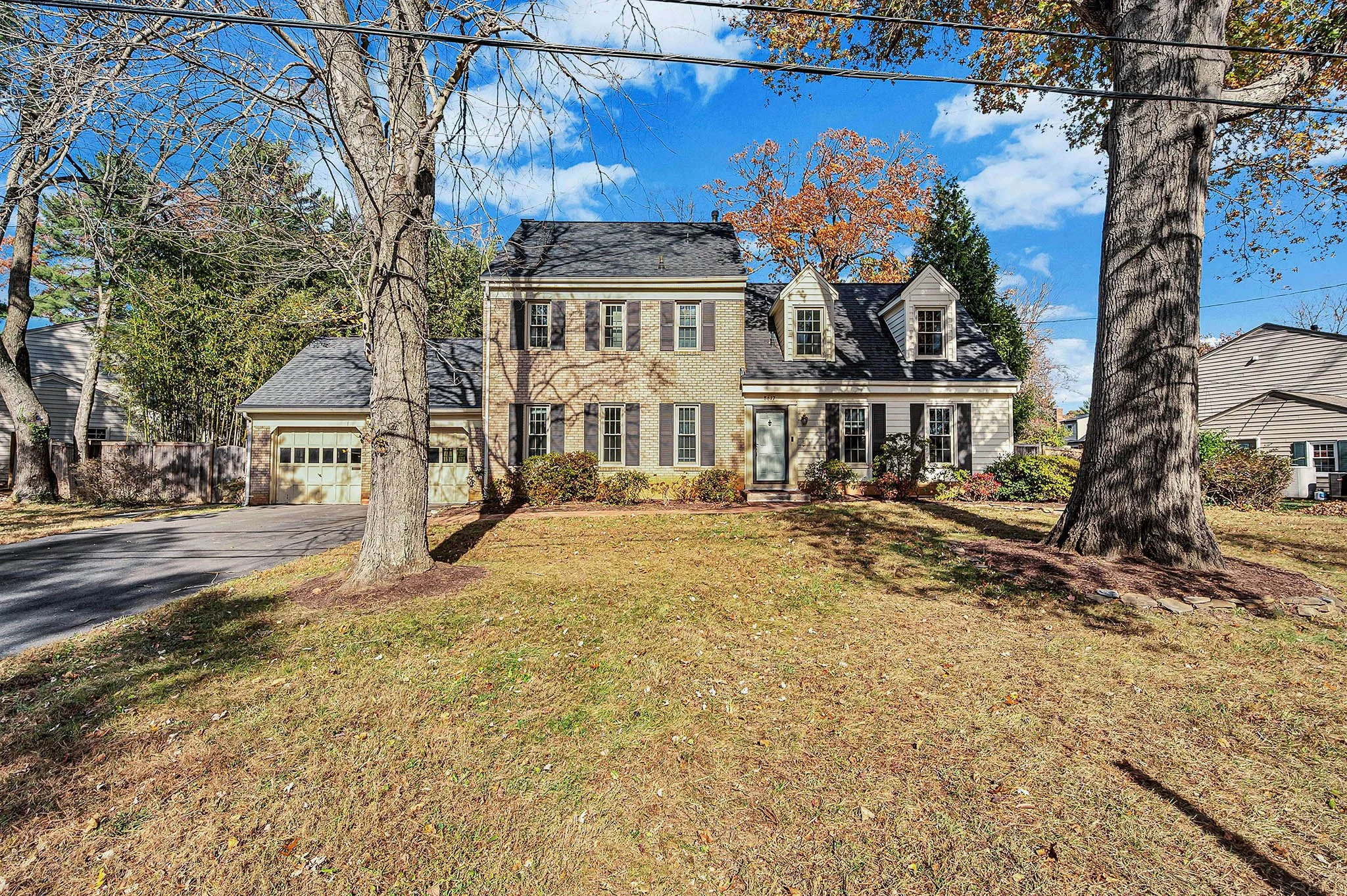 A two-story house with a brick facade and a black shingle roof, surrounded by a lawn with large trees and shrubs, under a bright blue sky with some clouds.