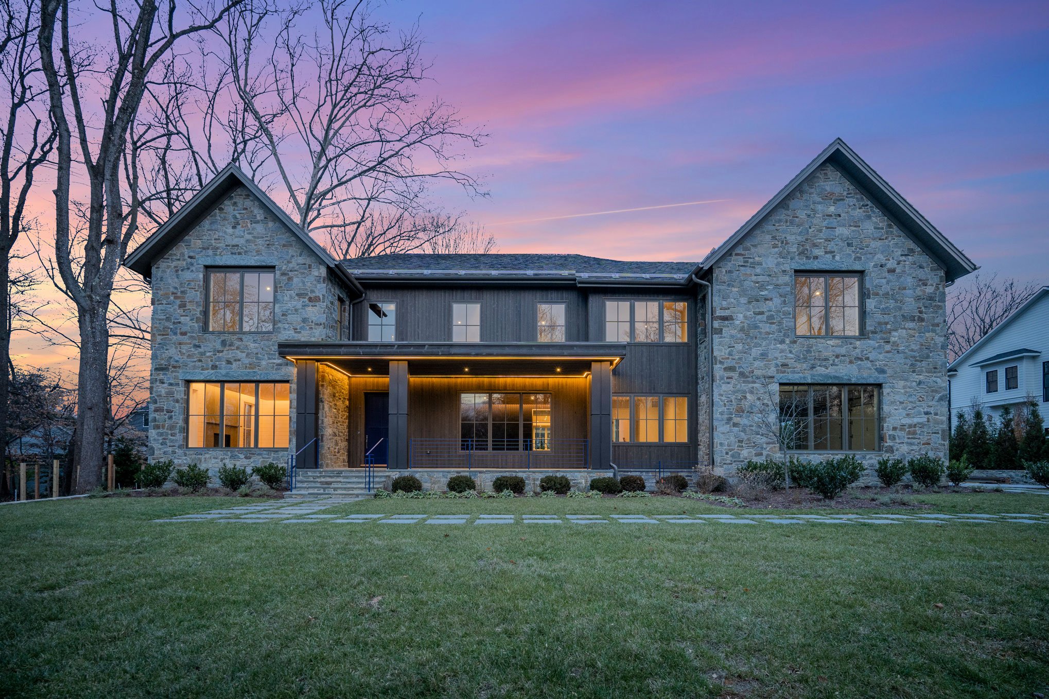 Modern house with stone and dark wood exterior, lit porch, large windows, and a spacious green yard during sunset.