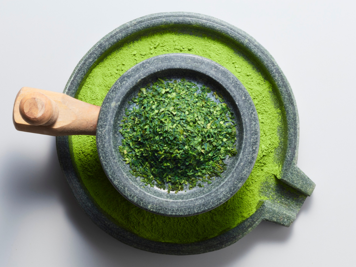 Top-down view of a gray stone mortar and pestle containing dried green matcha powder and chopped fresh matcha leaves, with a large matching stone bowl with remaining matcha powder around it.