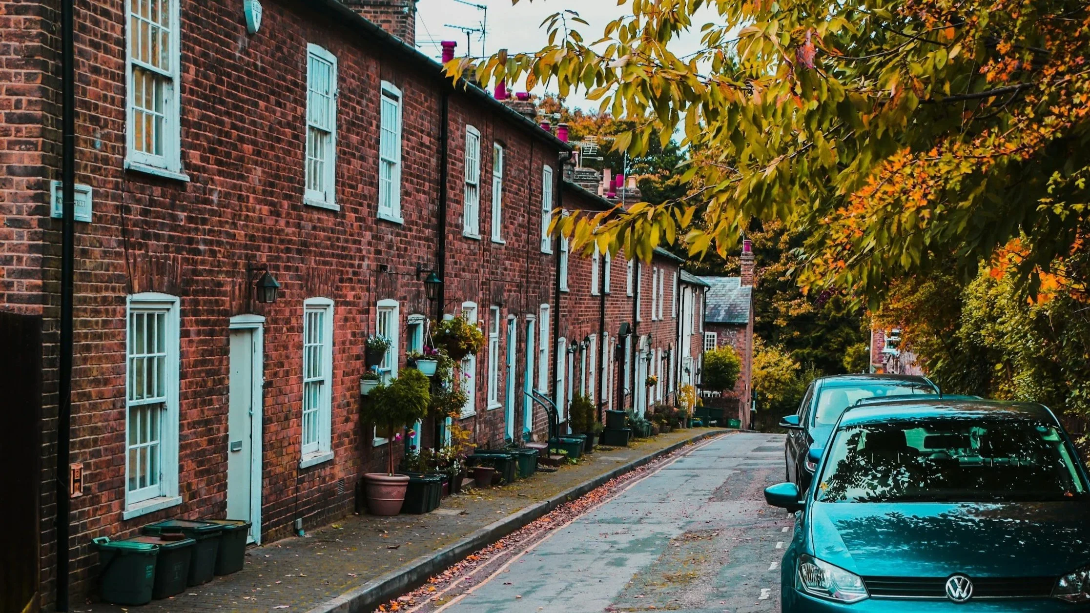A quiet residential street with red brick row houses and a sidewalk lined with potted plants and hanging flowers, surrounded by trees with autumn-colored leaves and parked cars.