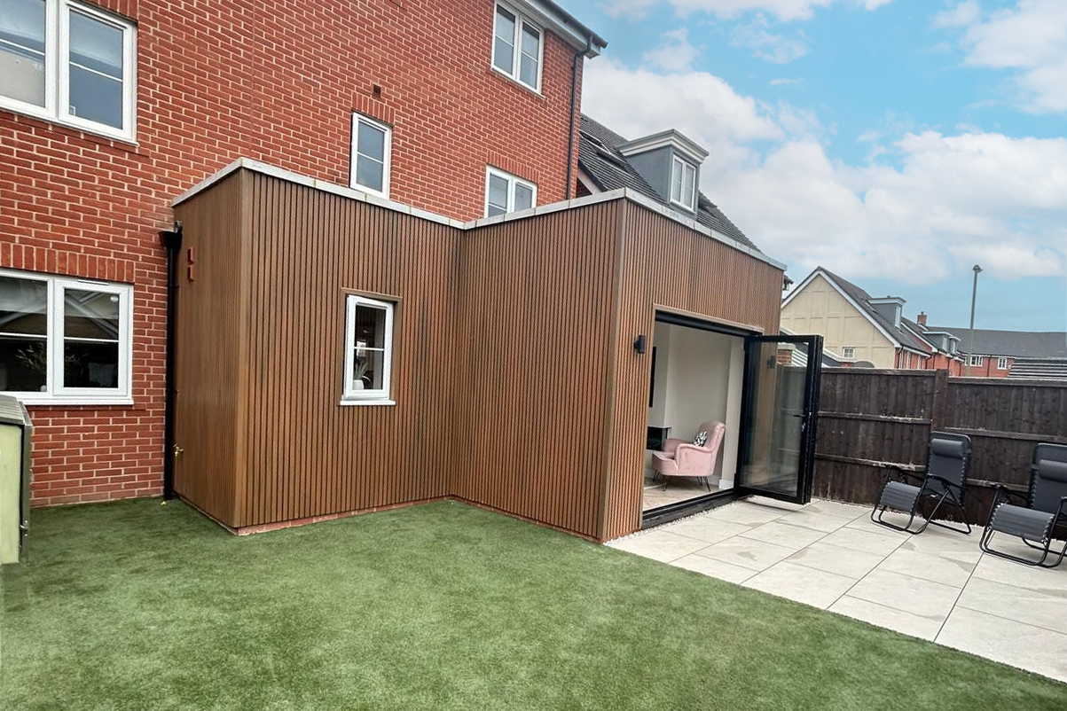 Backyard with small wooden extension attached to brick house, large sliding glass door open to pink armchair inside, patio area with lounge chairs, fenced yard under partly cloudy sky.