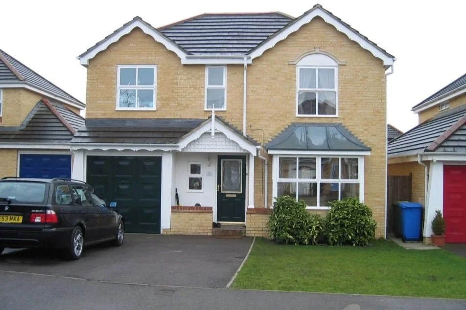A two-story yellow brick house with a black front door, a small porch, and a bay window with a tin roof, with a driveway and a black car parked in front. There are bushes, a blue trash bin, and a small potted plant near the house.