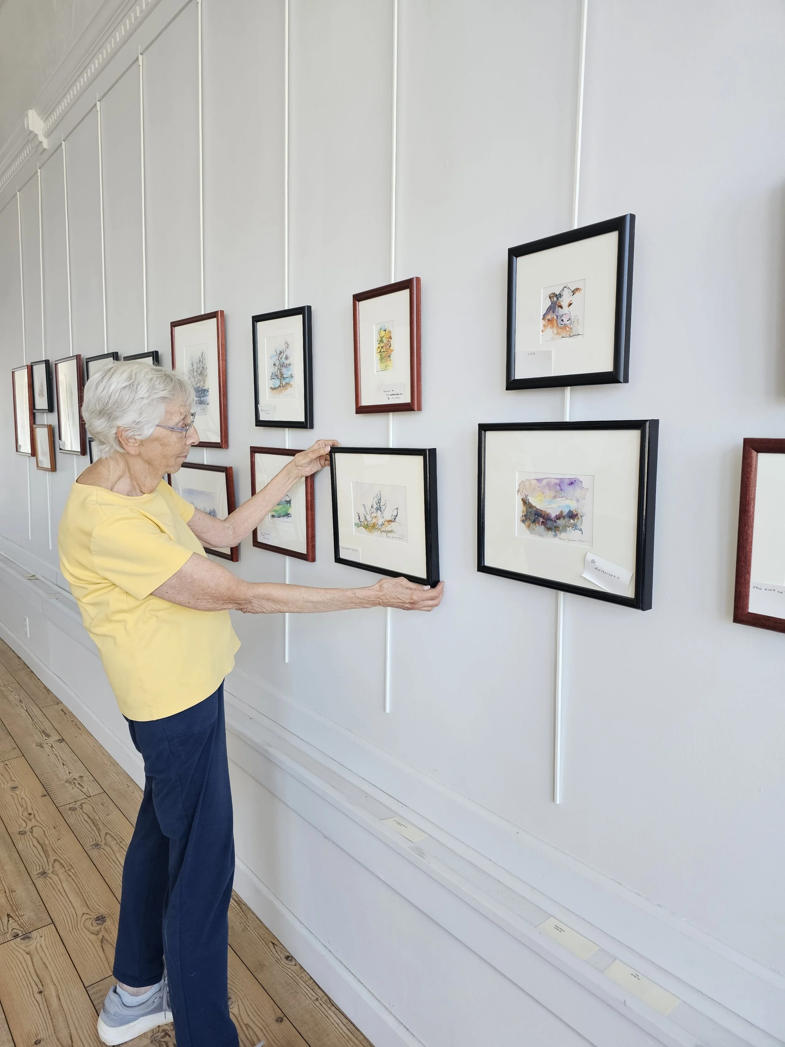 An elderly woman in a yellow shirt and navy pants looking at framed artwork displayed on a white gallery wall.