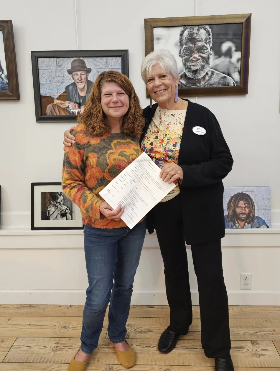 Two women smiling and holding a certificate, standing in front of a wall with framed black-and-white portraits of people.