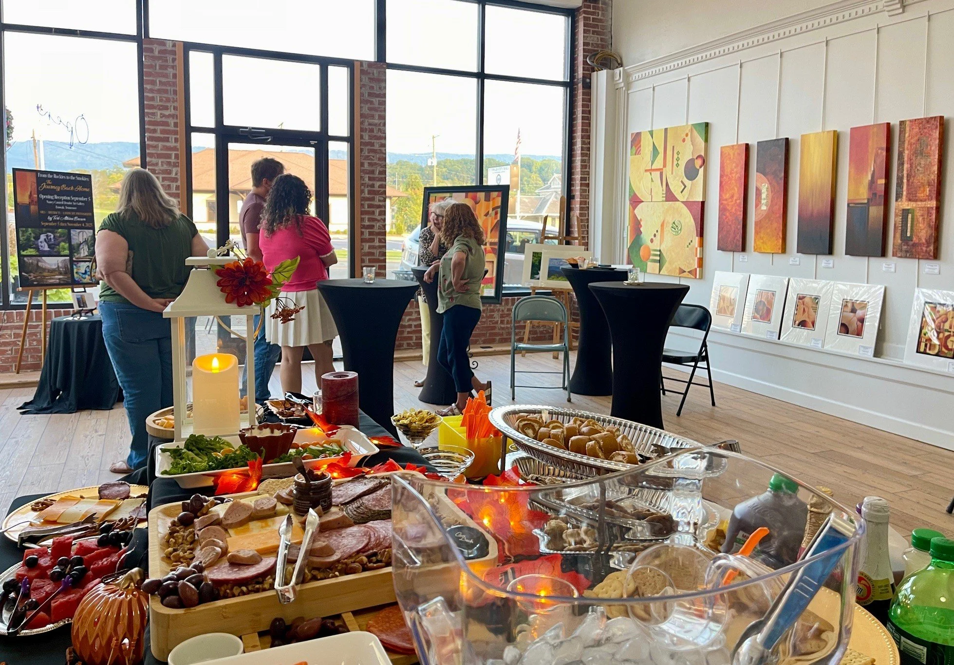 People at an art gallery event with colorful paintings on the wall, high tables, and a spread of food and drinks on a table in the foreground.